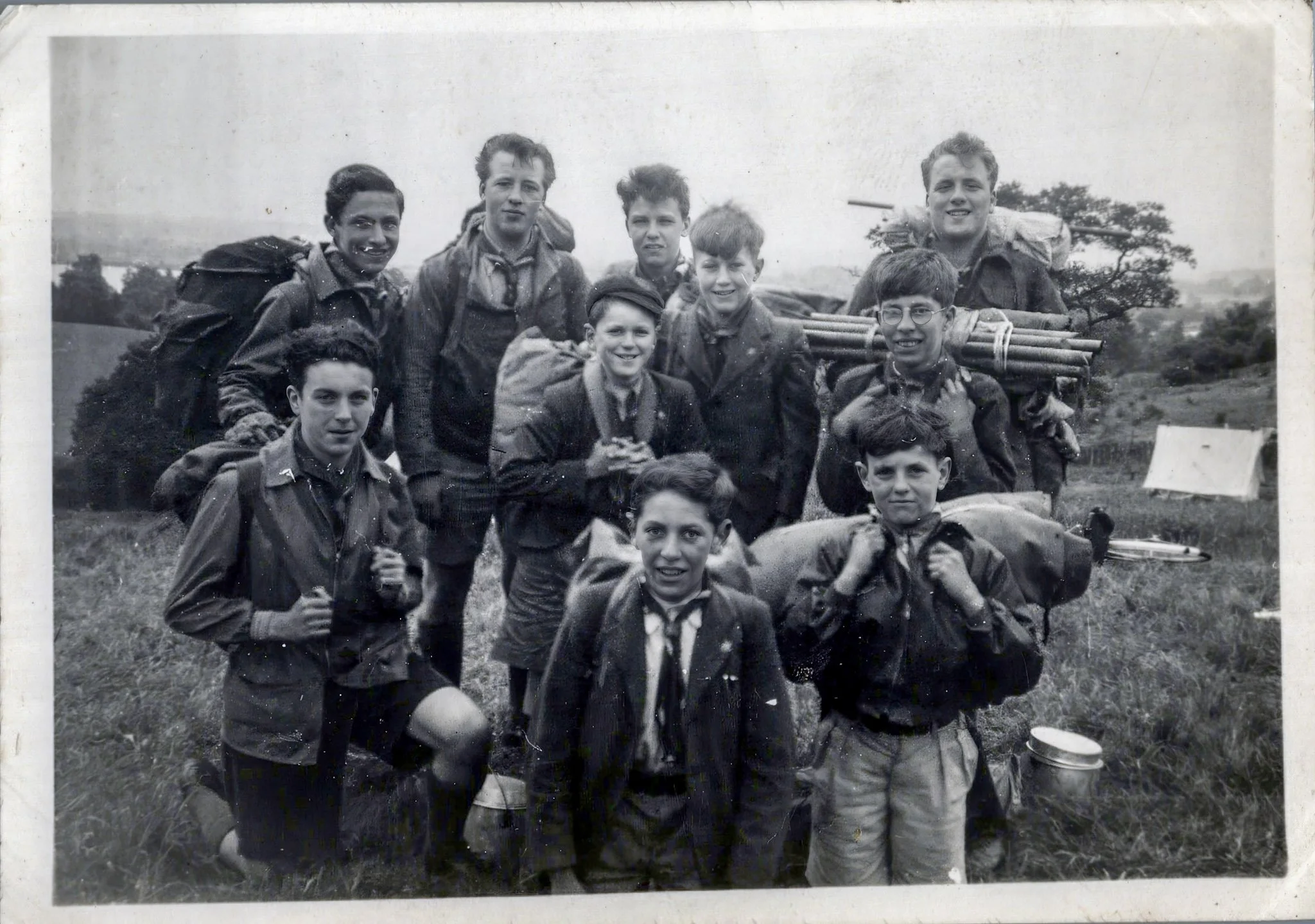 A black-and-white photograph of twelve young boys in outdoor camping gear, carrying backpacks and equipment, standing in a grassy field with trees and open land in the background.