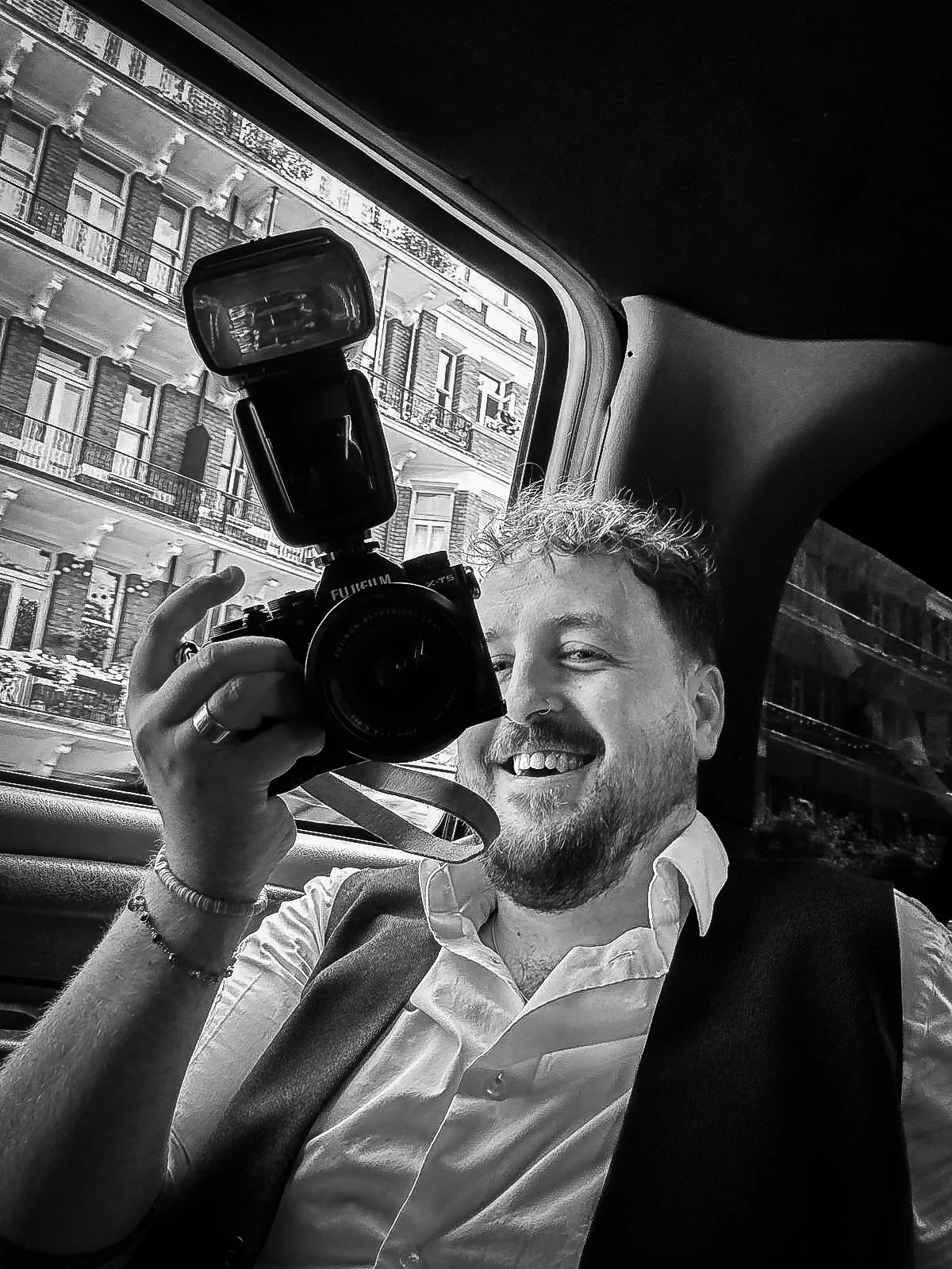 Black and white photo of a smiling person holding a camera with a flash in a car, with a view of a building outside the window.