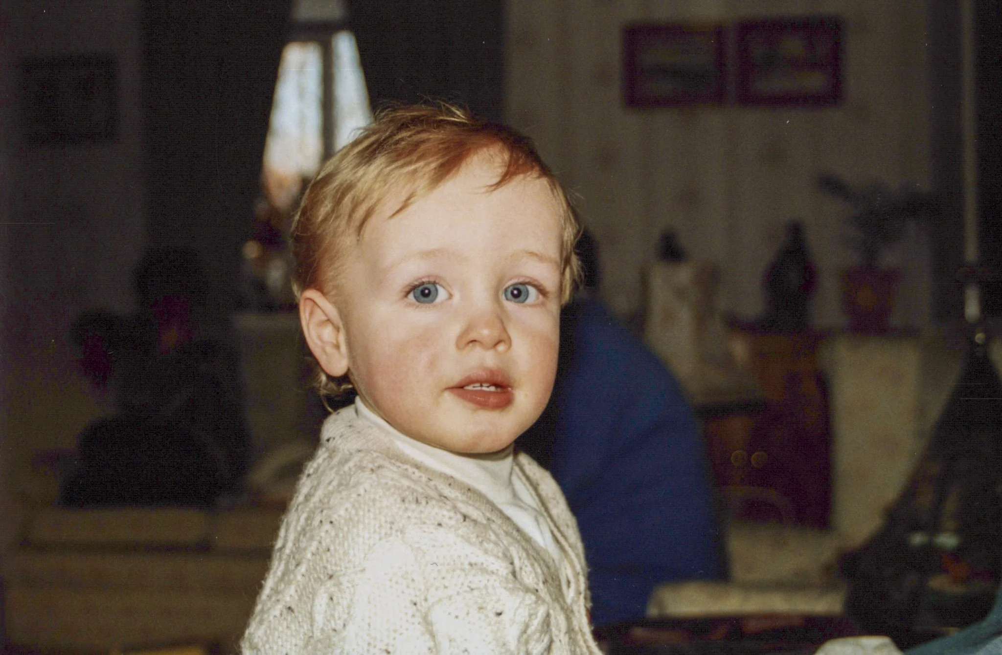 A young boy with red hair and blue eyes looking at the camera, wearing a cream-colored knitted sweater, inside a home with a window, artwork, and decorative items in the background.