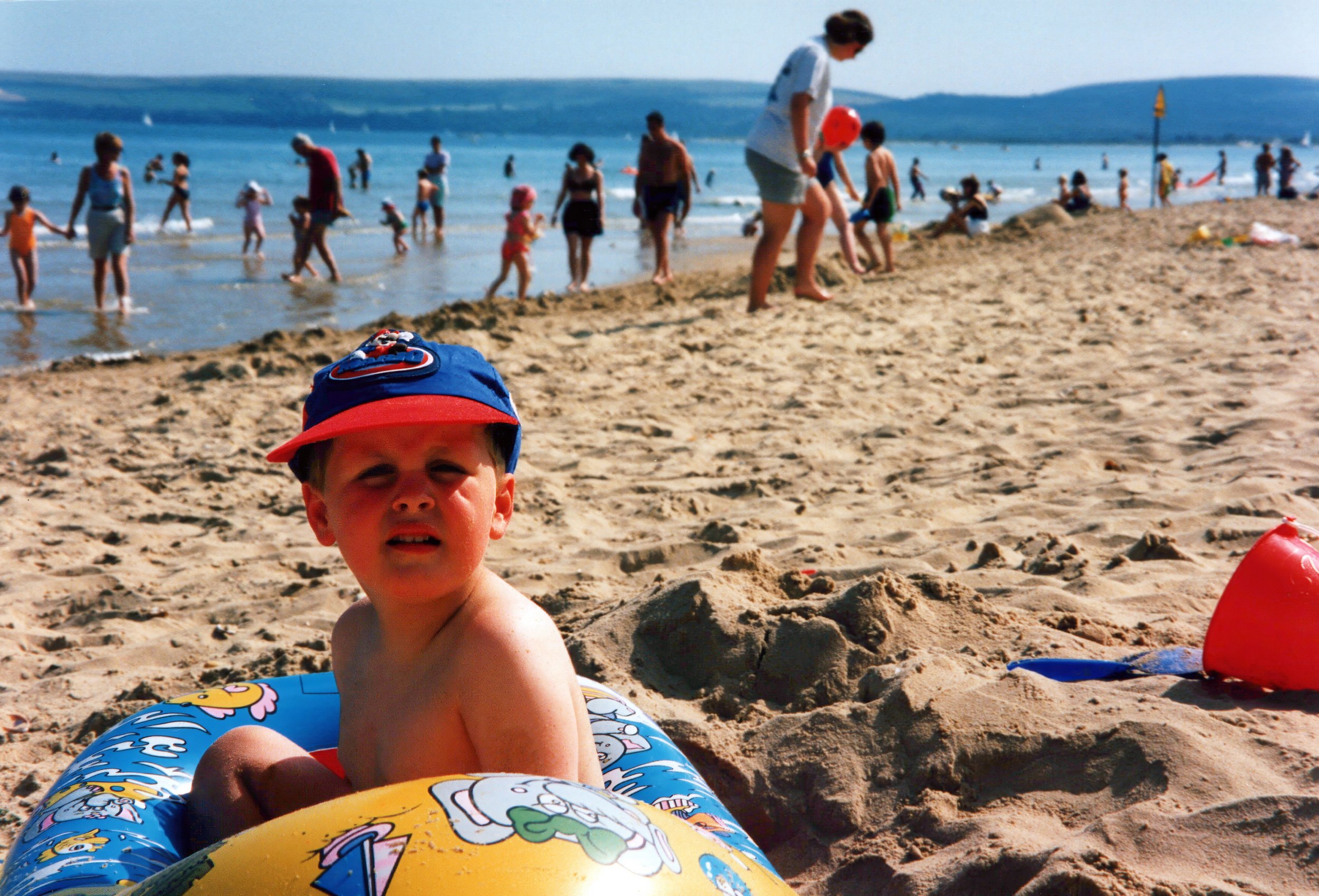Child in inflatable ring on sandy beach with people and ocean in background.