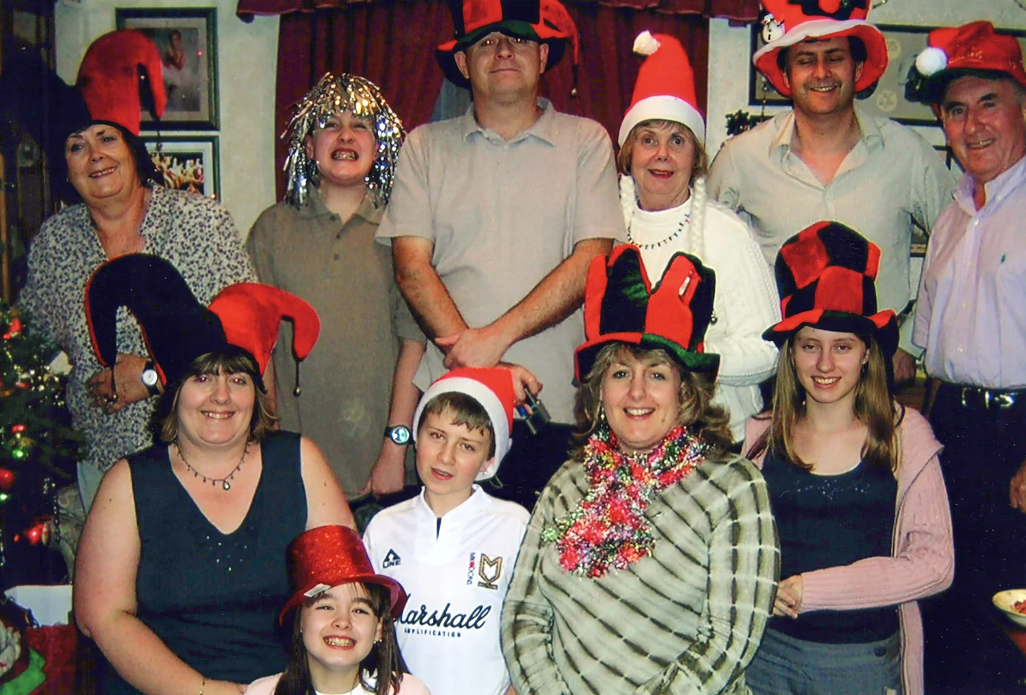 Group of people celebrating Christmas, wearing Santa hats and festive accessories, standing indoors near Christmas decorations and a Christmas tree.