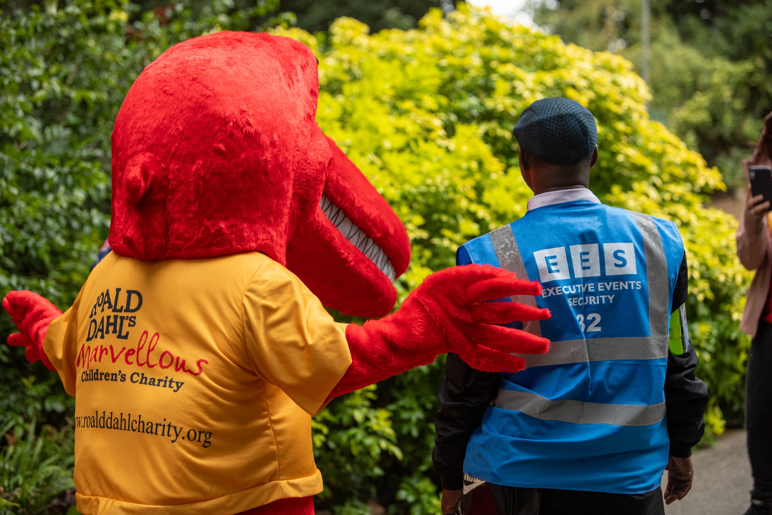 A person in a red mascot costume with a yellow shirt featuring Roald Dahl's charity logo approaches a security guard in a blue vest. They are outdoors near green foliage.