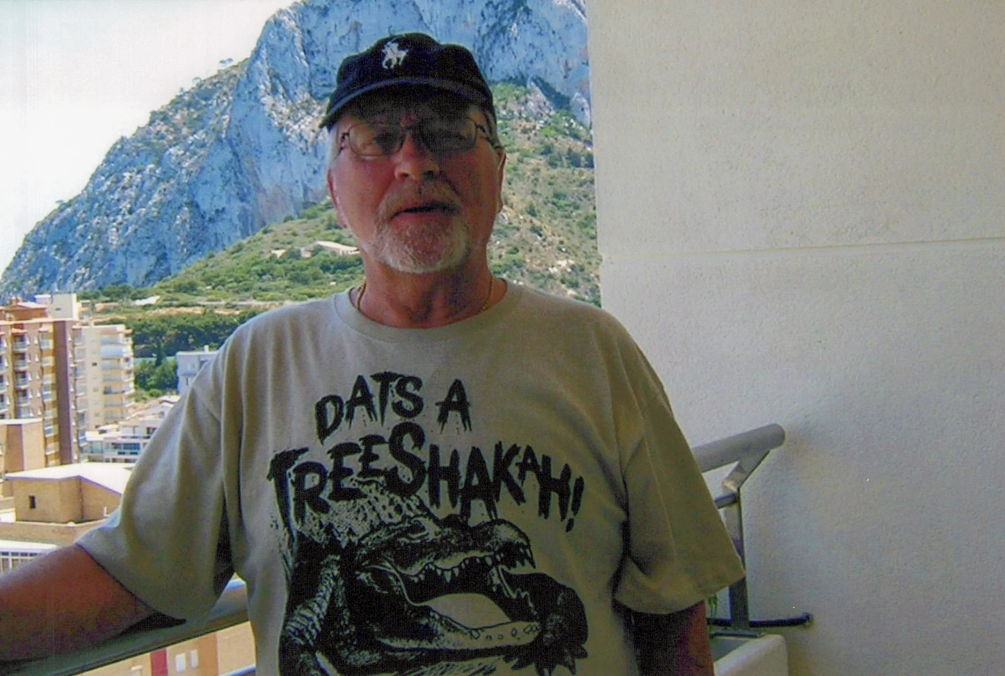 An older man wearing a beige T-shirt with a dinosaur graphic and the text 'DATS A TRESHAKH!' standing on a balcony with a cityscape and mountain in the background.
