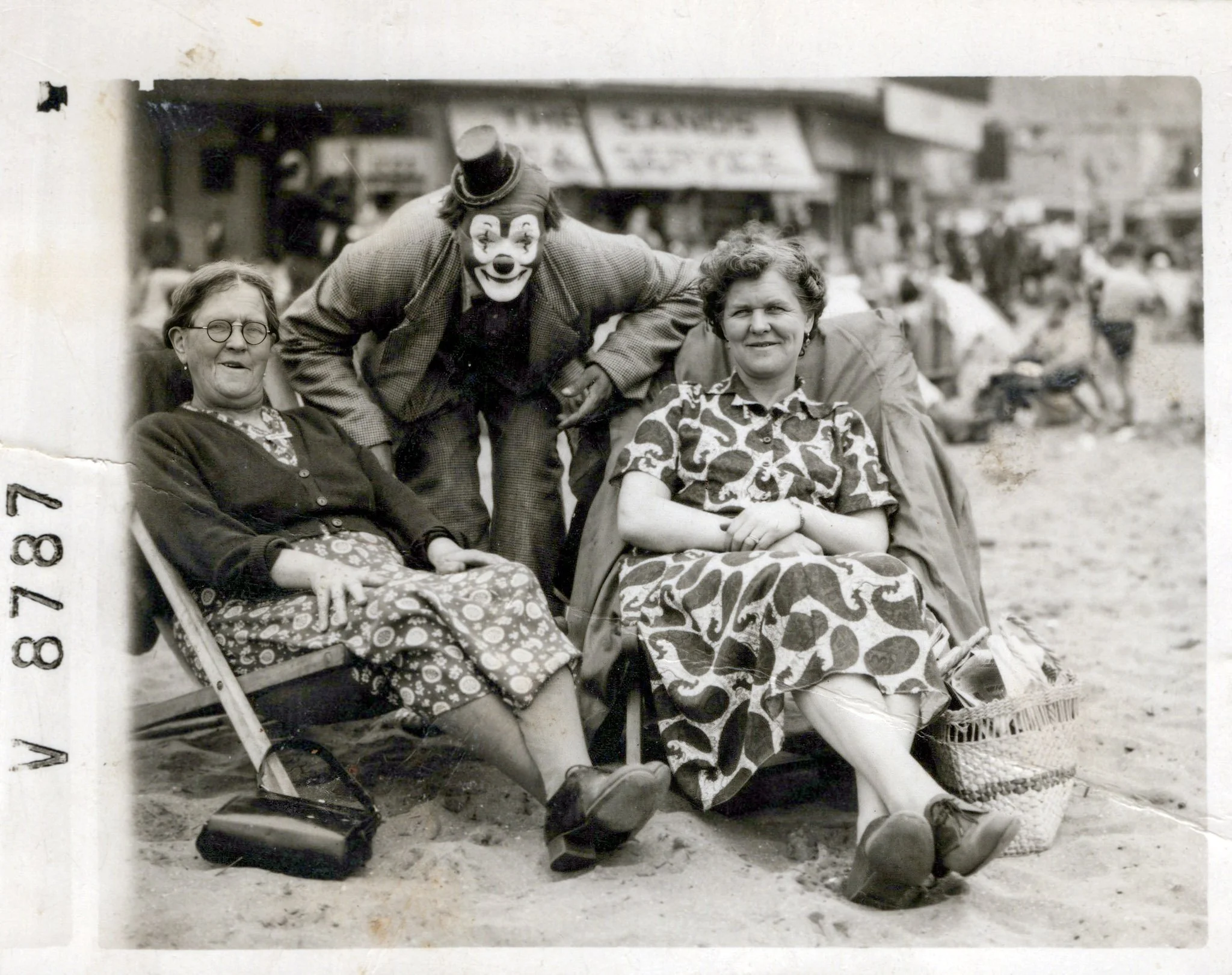 Black and white photo of two women sitting on beach chairs, with a person dressed as a clown standing behind them. The women are wearing patterned dresses and the clown is in costume with a painted face, hat, and blazer. Background shows a busy beach scene with people and a boardwalk.