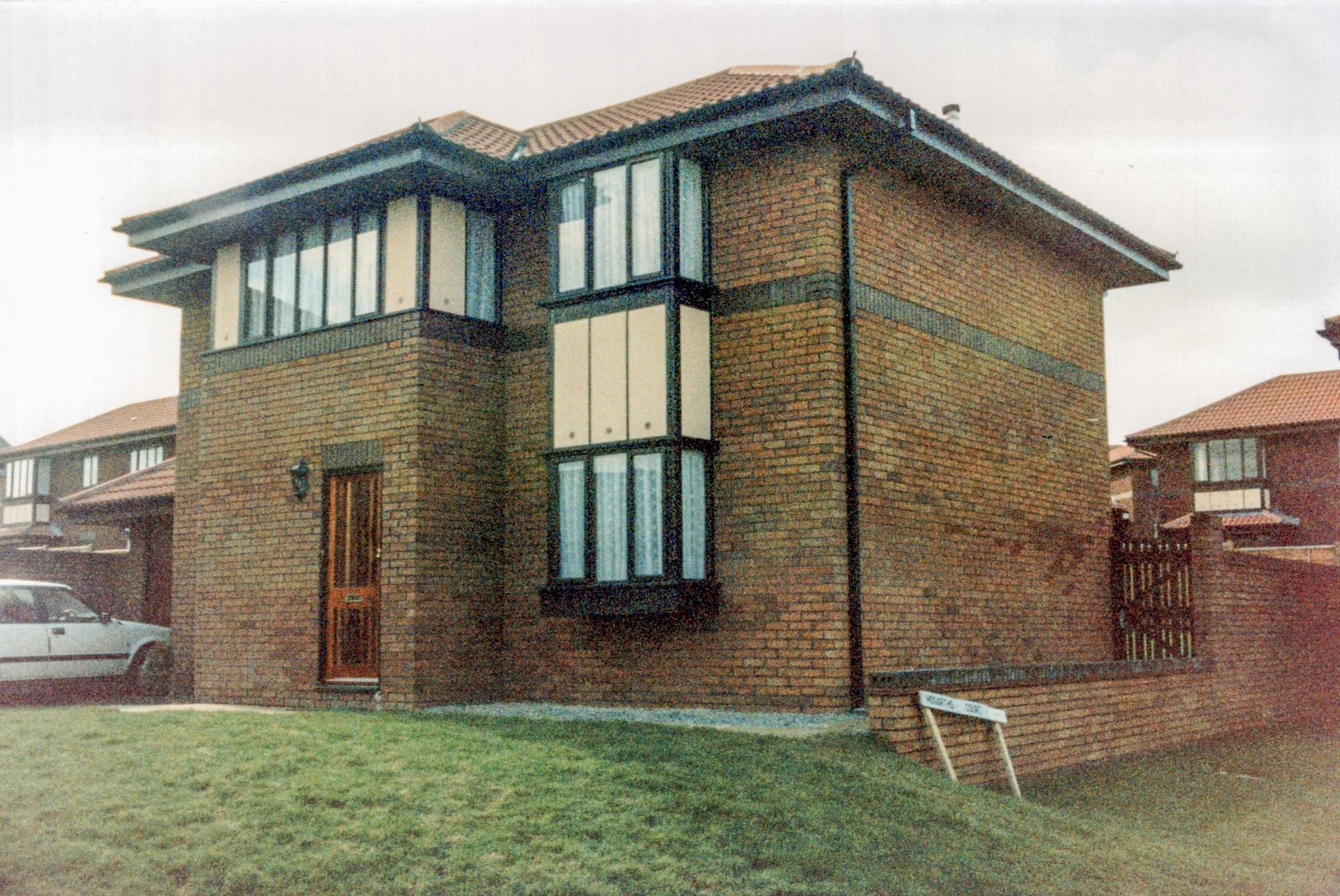 Two-story brick house with a wooden front door, large front windows, and a small front lawn, with neighboring brick houses visible in the background.