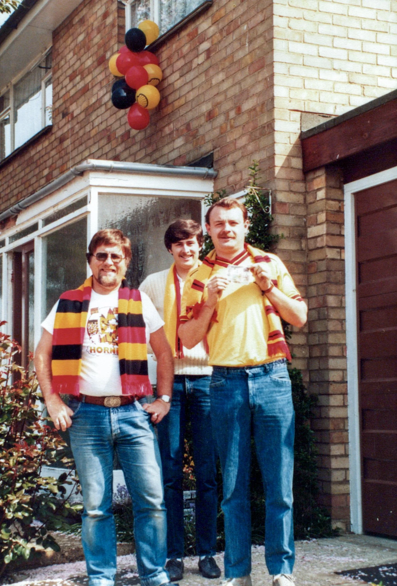 Three men standing outside a brick house decorated with black, red, and yellow balloons, wearing football scarves and jerseys, celebrating a soccer event.