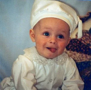 A smiling baby wearing a cream-colored hat and a white outfit, sitting indoors.