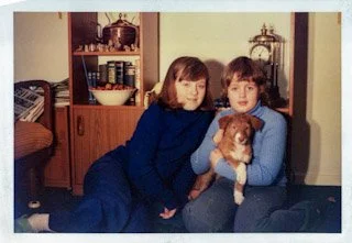 Two women sitting on the floor of a living room, holding a small dog, with a wooden cabinet and decorative items in the background.