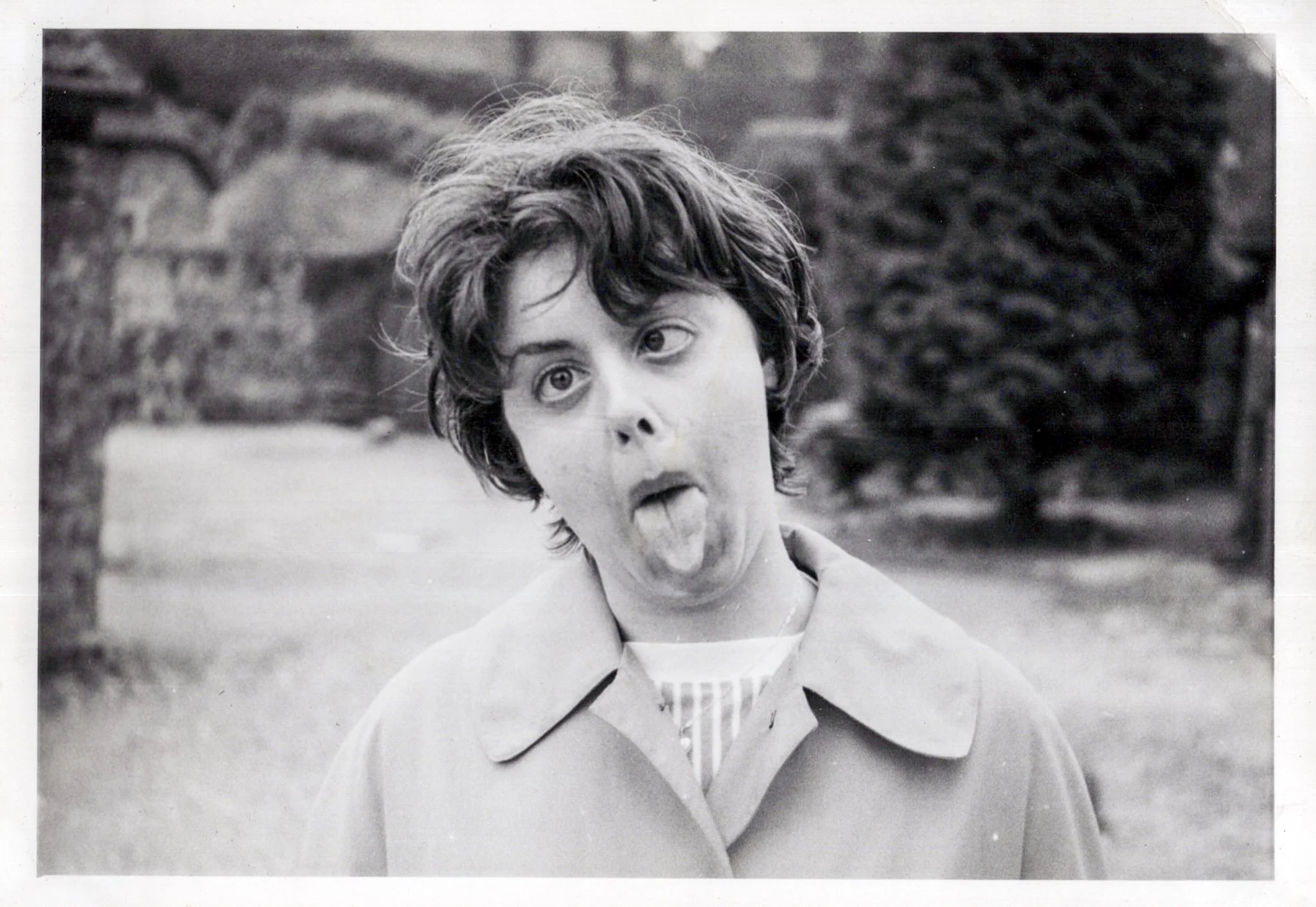 Black and white photograph of a woman making a silly face with her tongue out and eyes wide open. She has short, curly hair and is wearing a light-colored coat. The background shows some trees and a brick wall.