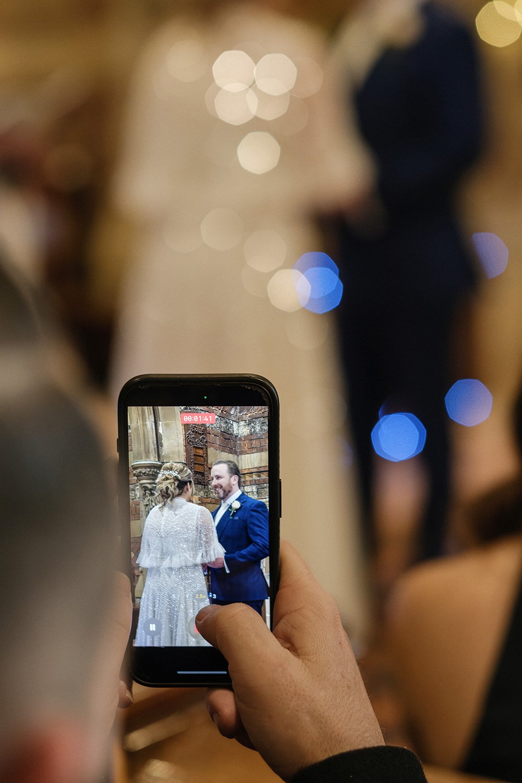 A person is recording or taking a photo of a wedding ceremony using a smartphone, with the couple standing in front of a brick and stone background.