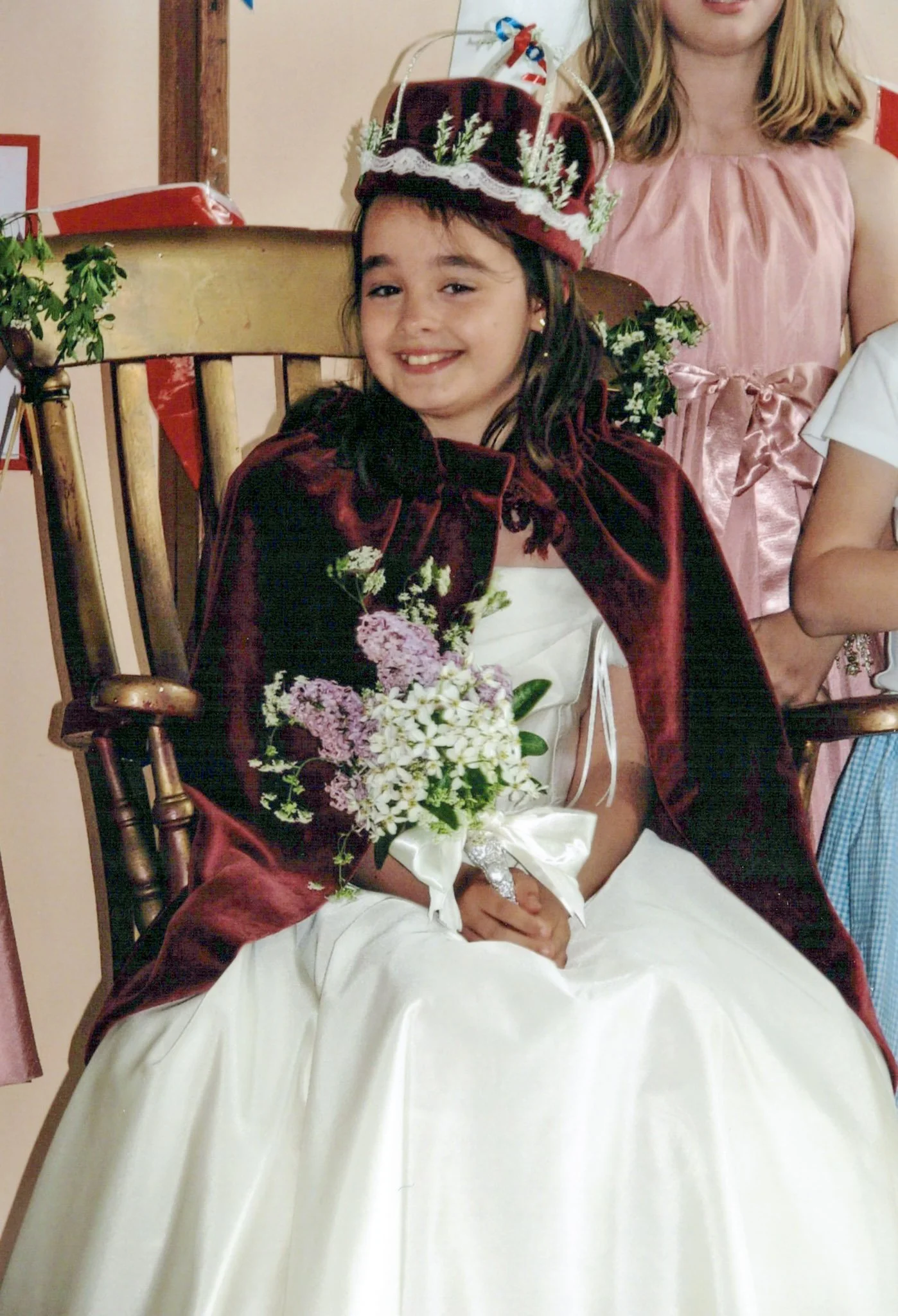 Young girl in a white dress holding a bouquet, wearing a maroon crown and cape, sitting on a wooden chair, smiling at a celebration or special event.
