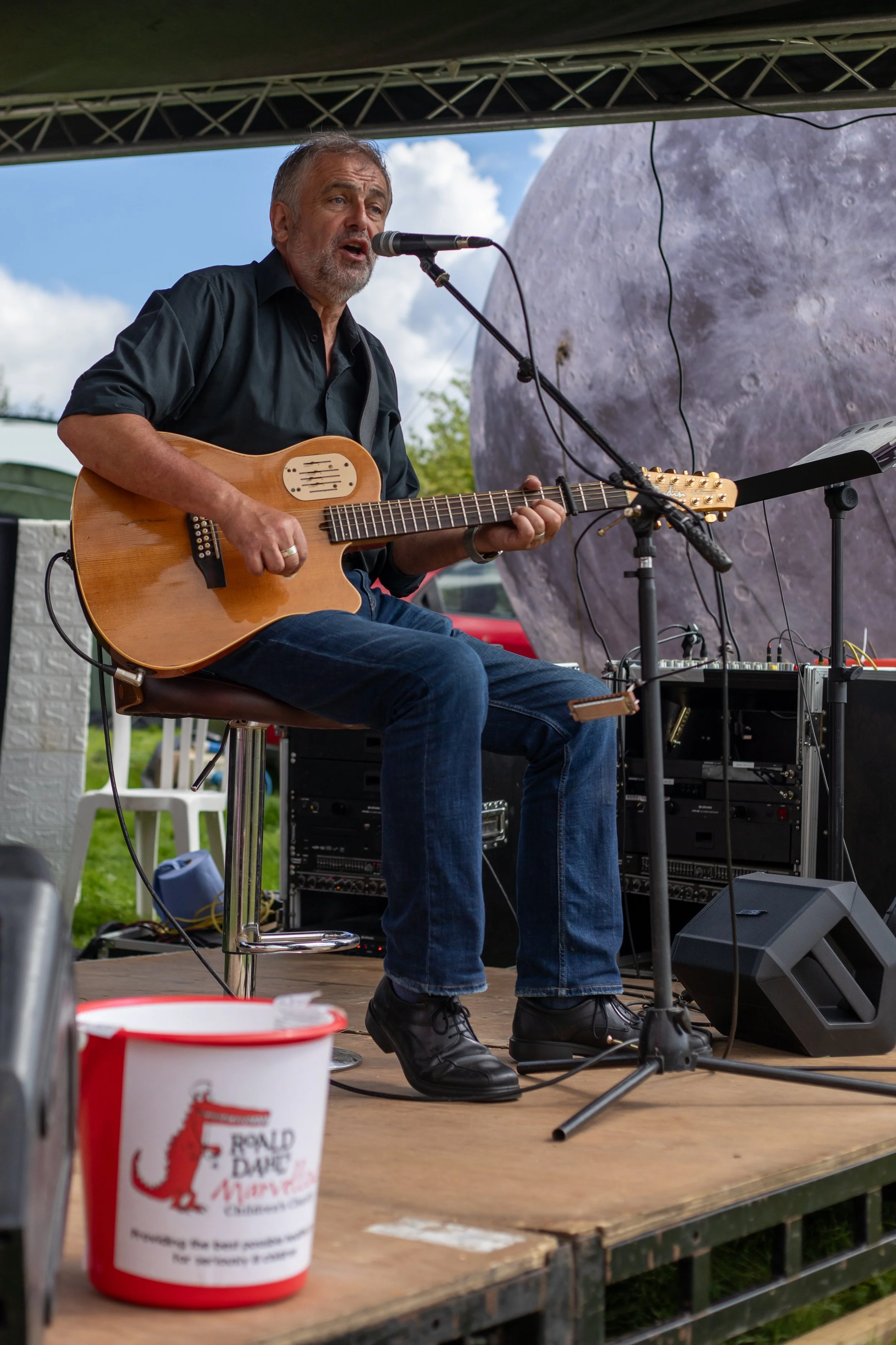 A man playing an acoustic guitar and singing into a microphone on an outdoor stage, with audio equipment visible and a charity donation bucket in the foreground.