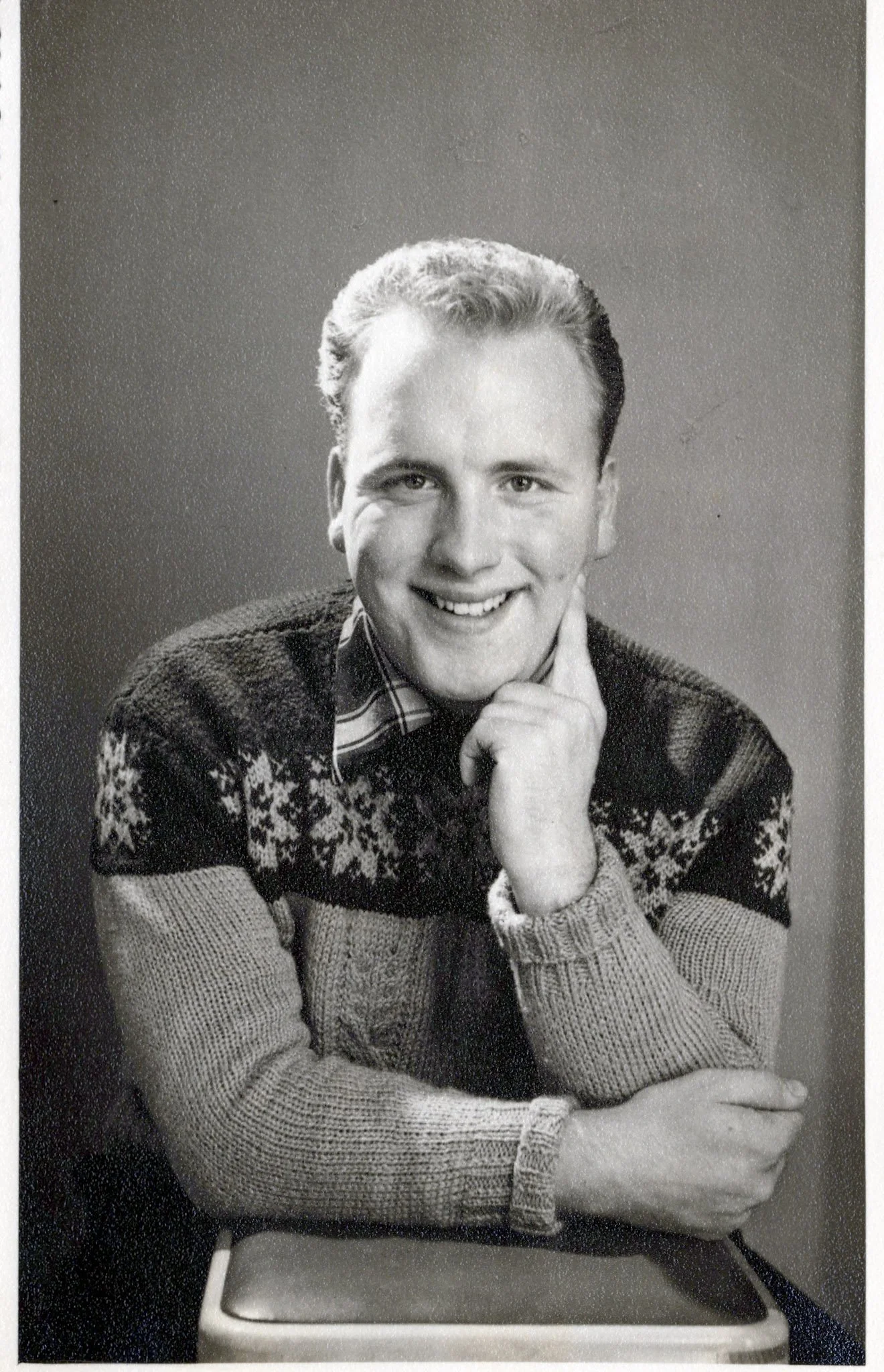 Black and white portrait of a smiling man with short hair, wearing a patterned sweater over a collared shirt, resting his chin on his hand, seated on a chair.