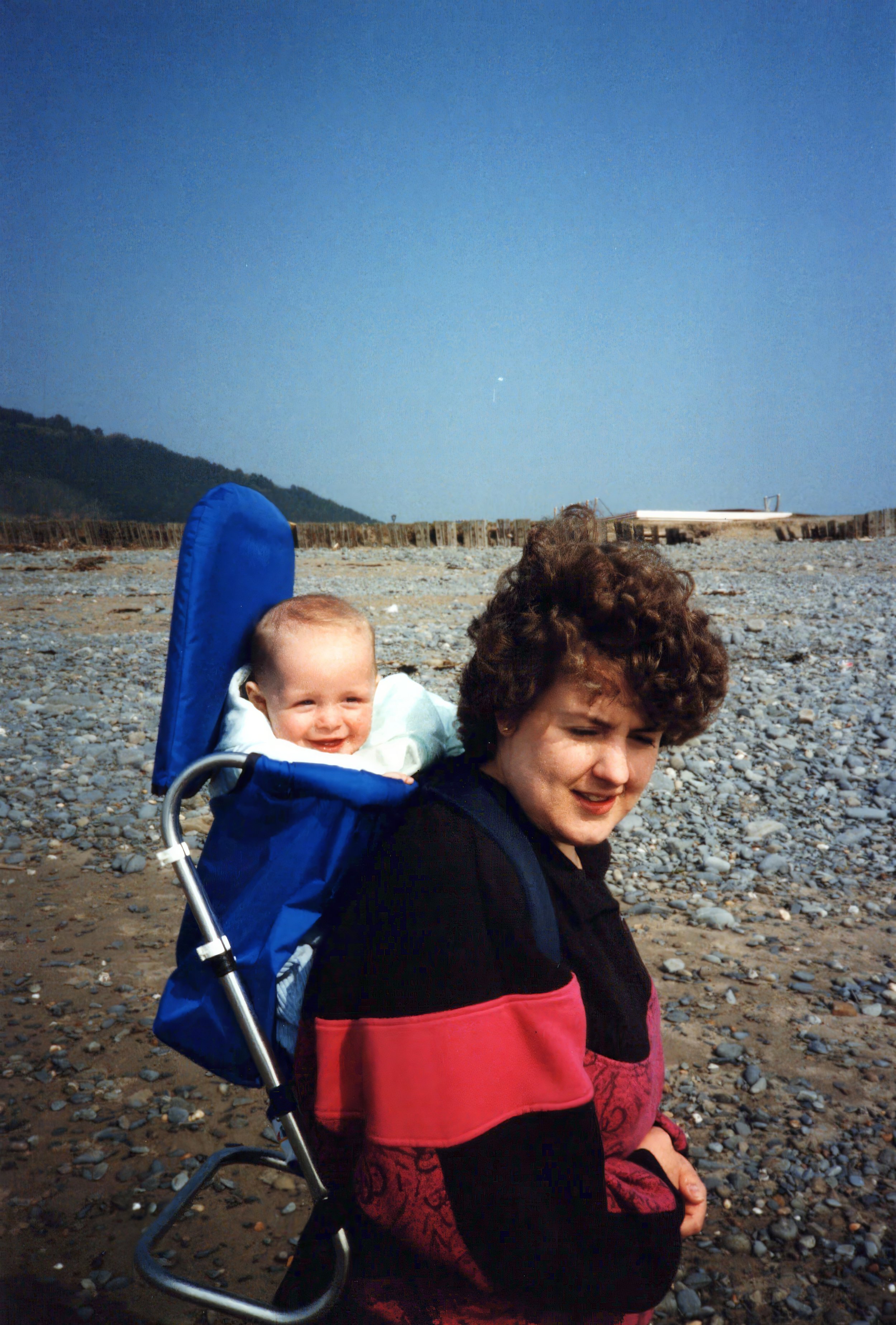 A person carrying a baby in a blue carrier on a rocky beach with hills and clear blue sky in the background.
