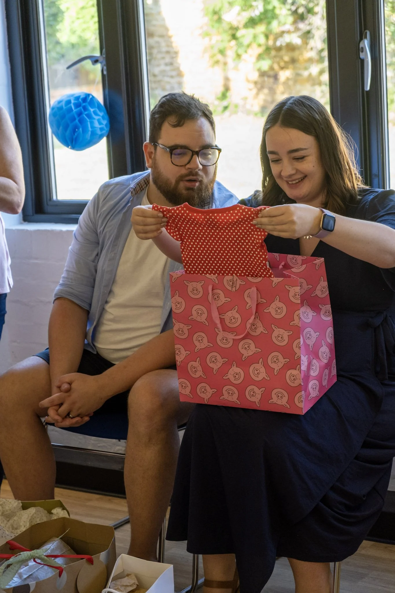 Two people sitting on a bench opening a pink gift bag with pig face designs, with a woman smiling and holding a red polka-dotted cloth.