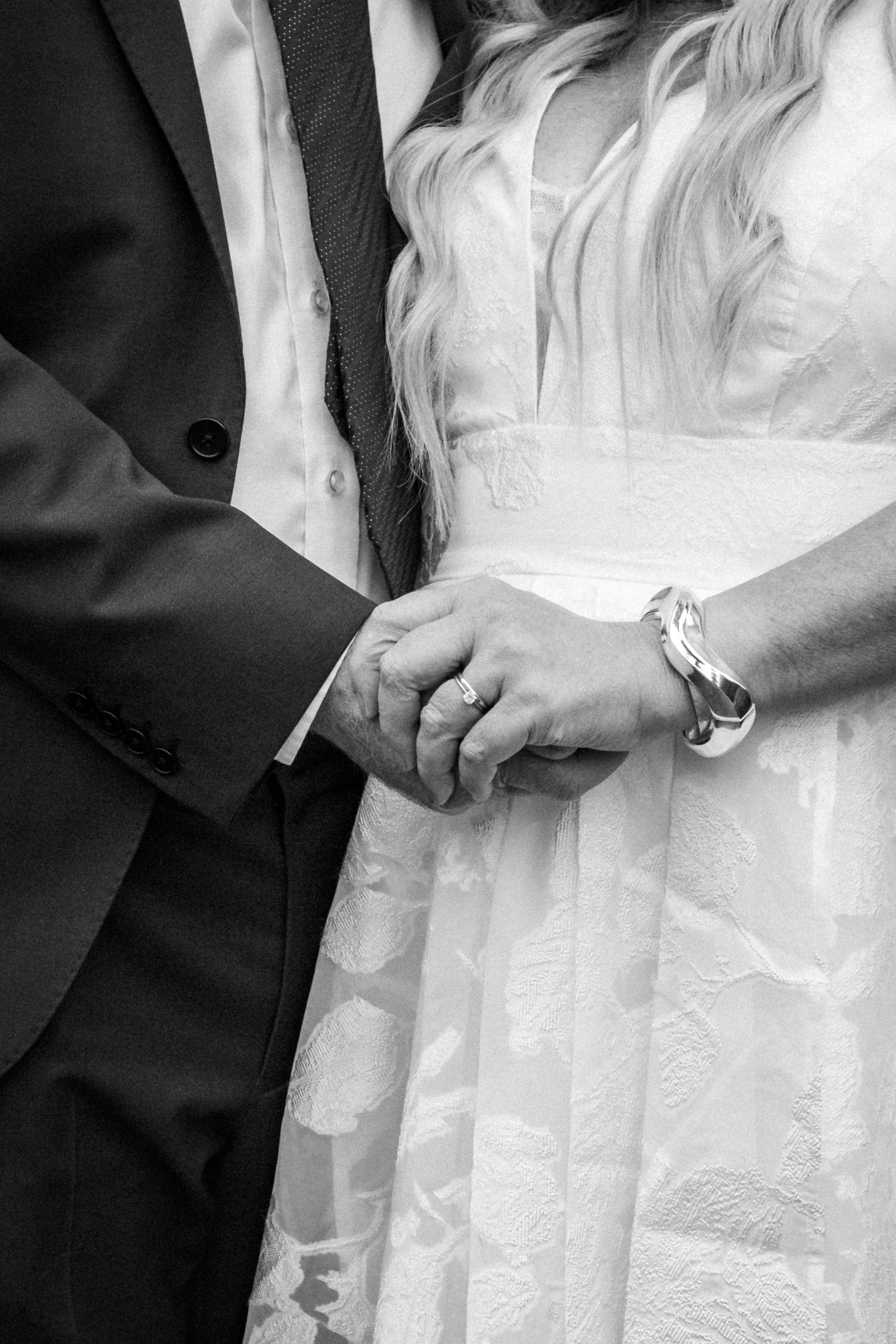 Close-up of a couple holding hands during a wedding ceremony, with the man wearing a suit and the woman in a wedding dress, both displaying wedding rings.