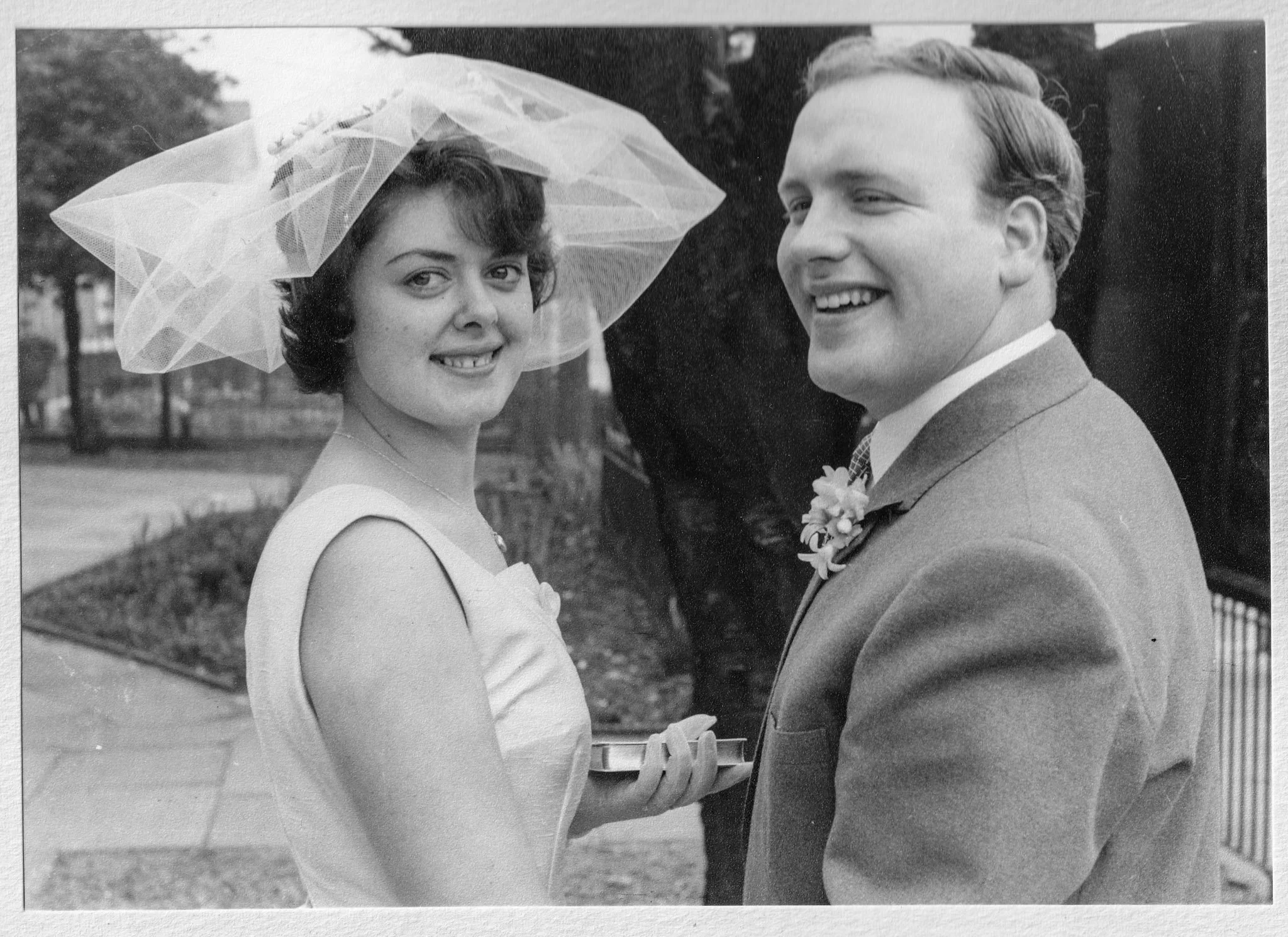 Black and white photo of a smiling woman in a hat and a man in a suit at a wedding, with the woman holding a tray.