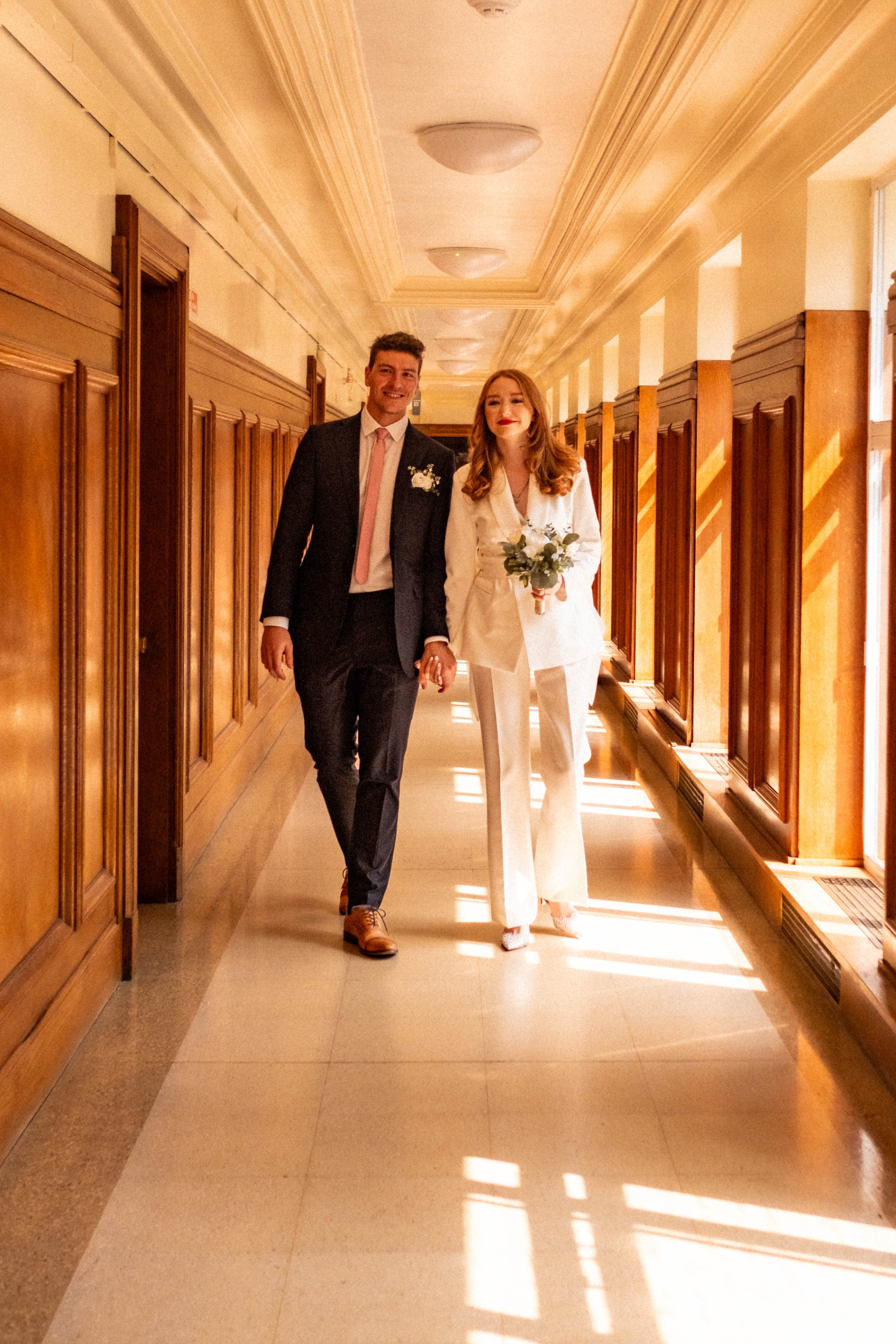 A couple, dressed in wedding attire, walking hand in hand through a sunlit hallway with wooden paneling, with the woman holding a bouquet of flowers.