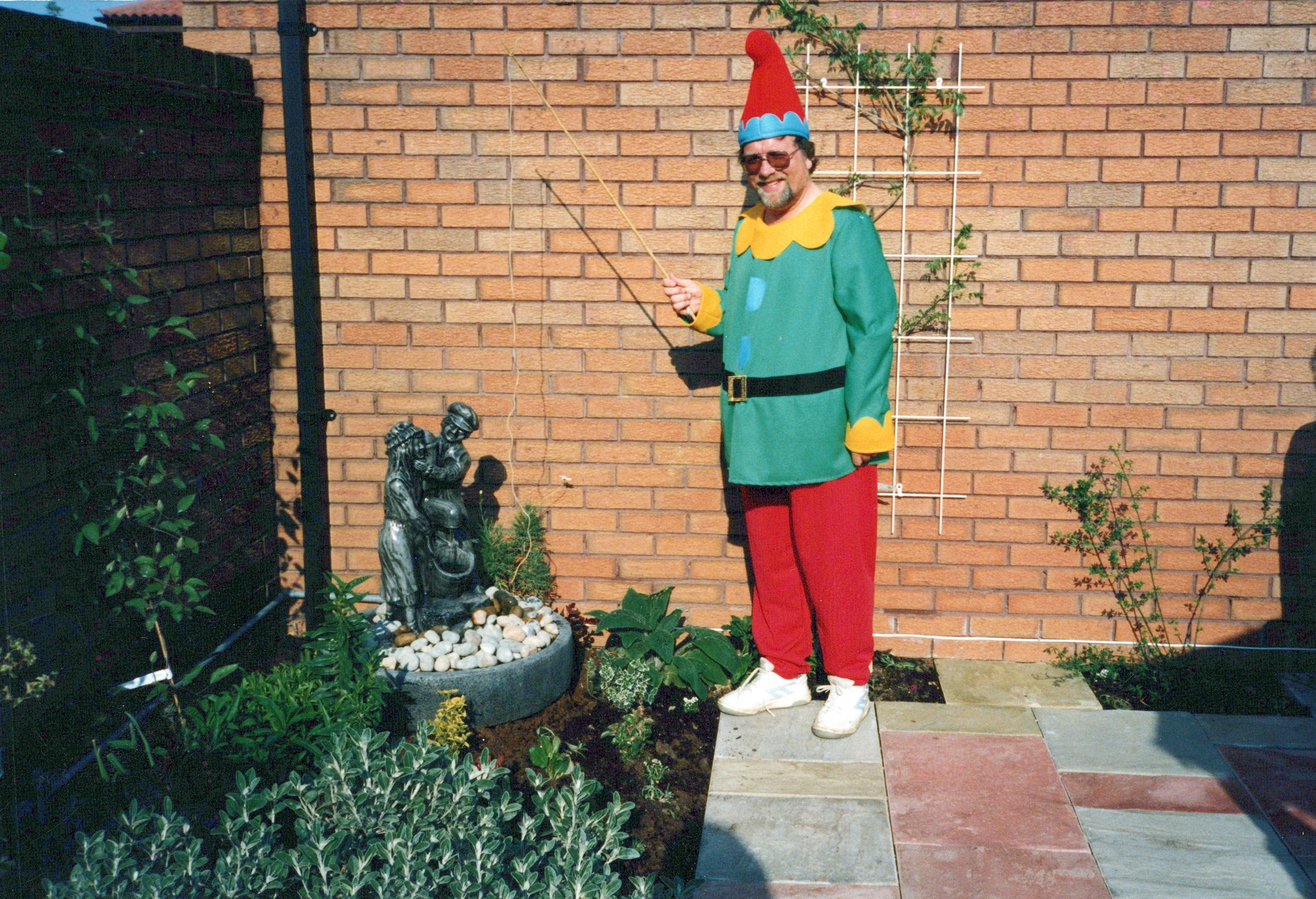 Man dressed as a garden gnome standing next to a garden fountain with two figures, in front of a brick wall and trellis.