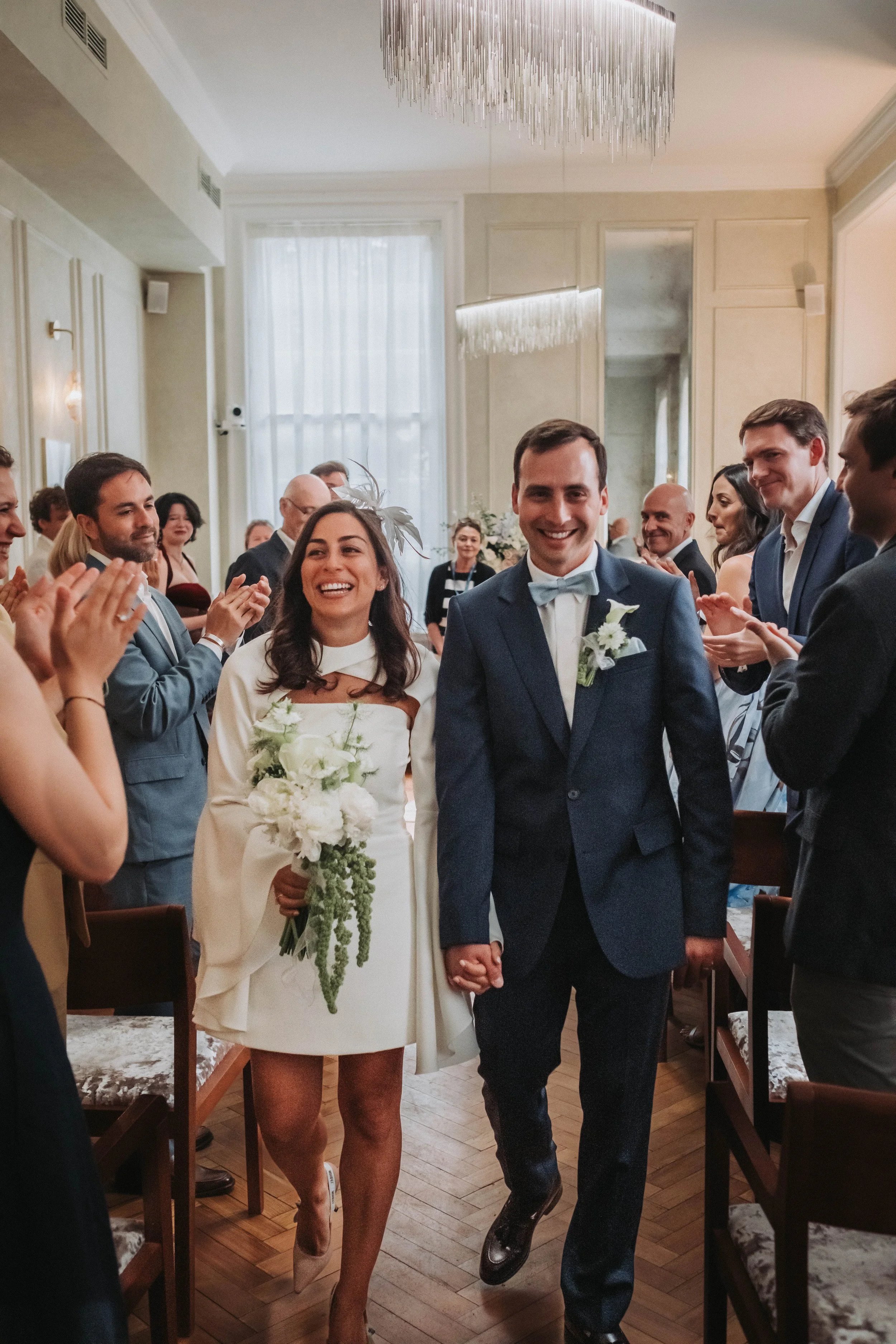 A happy bride and groom walking hand in hand through a crowd of smiling wedding guests in an elegant, well-lit room.
