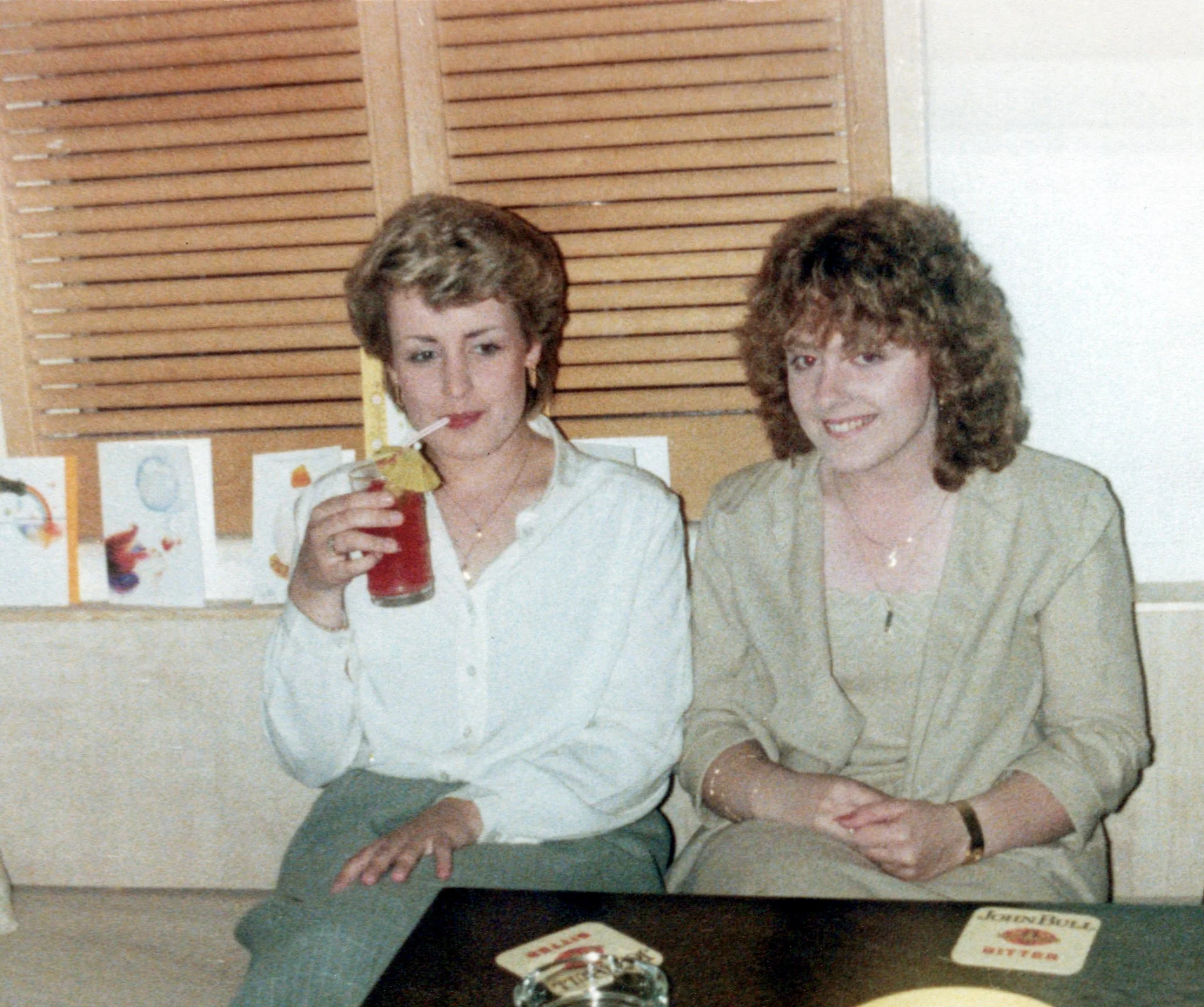 Two women sitting at a table, one drinking a red beverage with a lemon slice garnish, in an indoor setting with wooden paneling and greeting cards in the background.