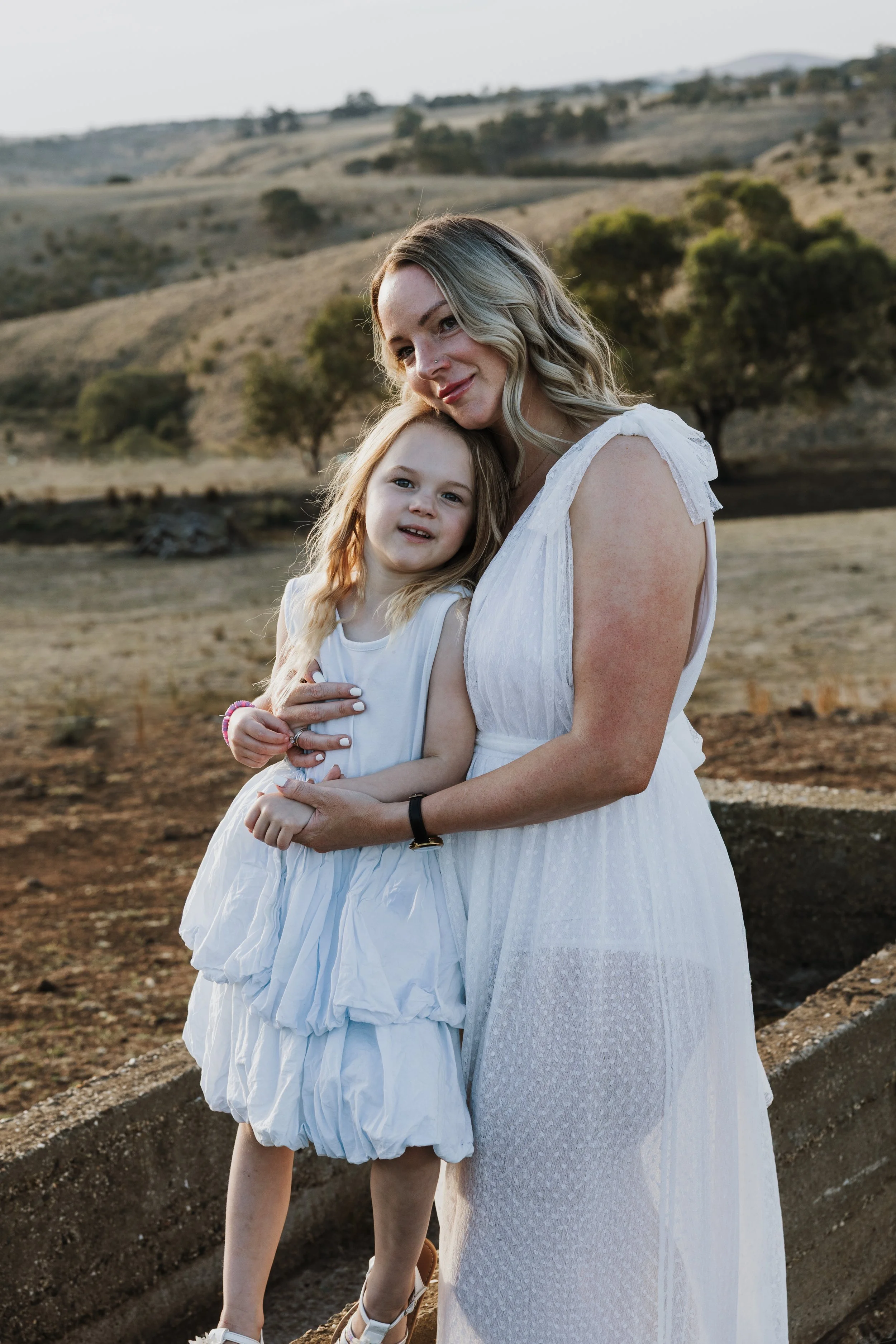Mother and daughter in white dresses embracing in a scenic outdoor setting with rolling hills and trees in the background.