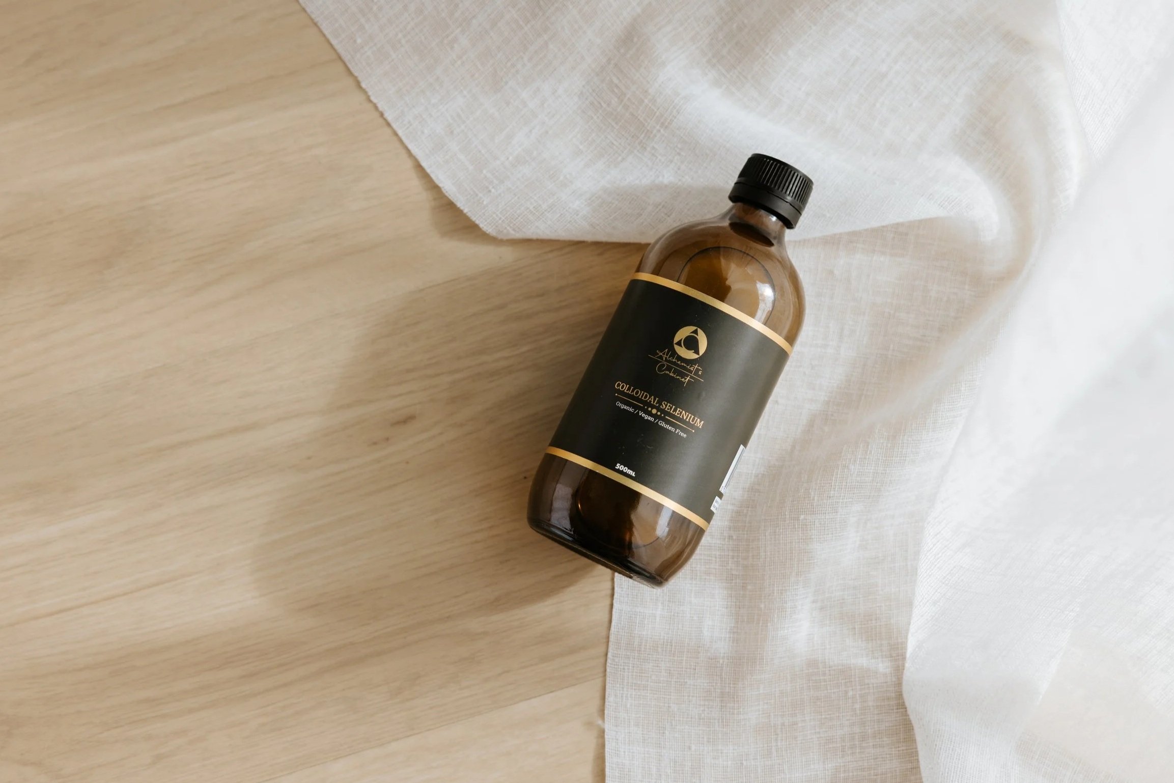 A brown plastic bottle with a black label and black cap, labeled "Colloidal Selenium" and indicating it is organic, vegan, and gluten-free, placed on a wooden surface with a white cloth underneath.