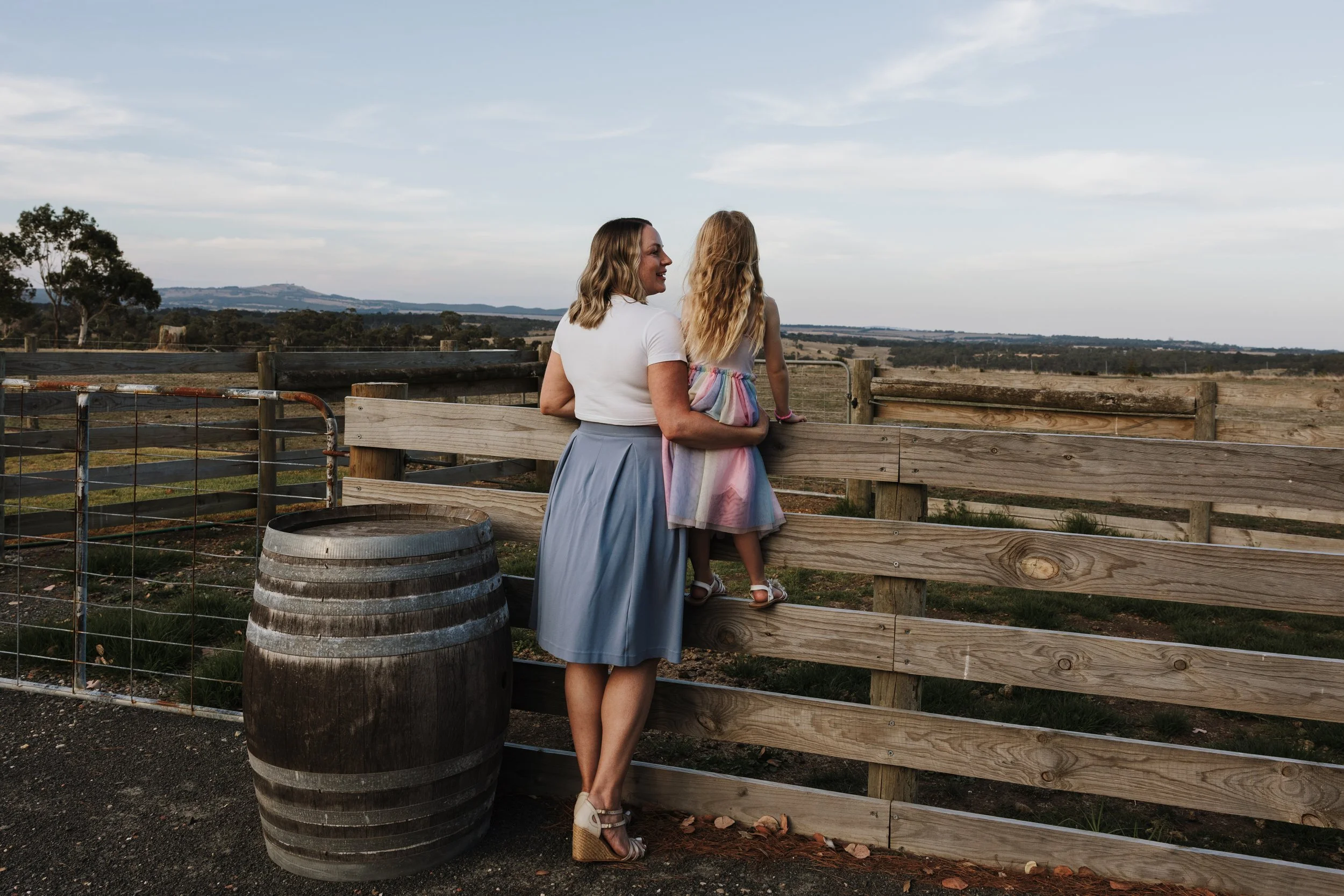 A woman and a young girl standing by a wooden fence, overlooking an open rural landscape with rolling hills and fields under a blue sky.