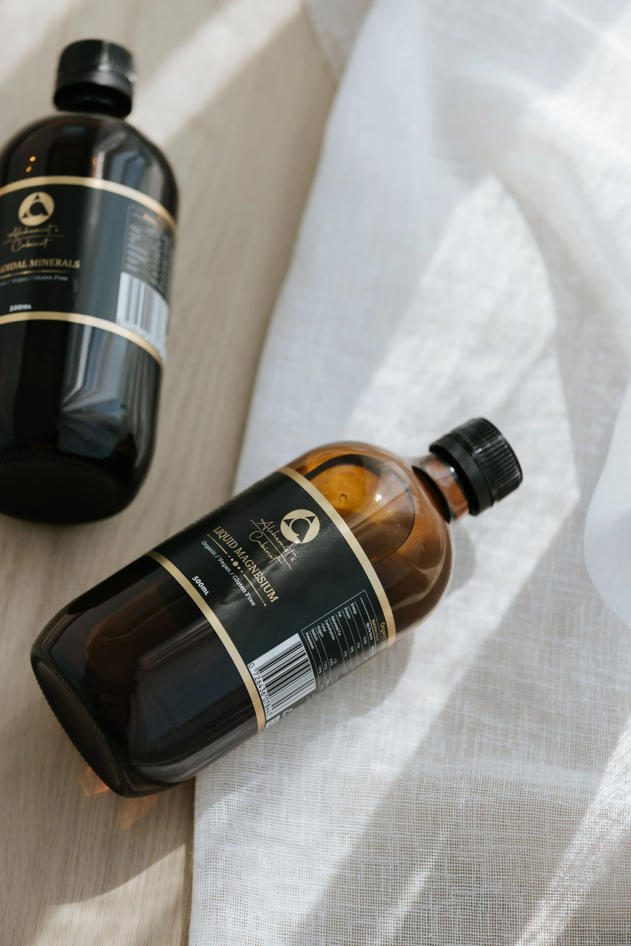 Two bottles of liquid magnesium with black labels and gold accents lying on a wooden surface and white fabric.