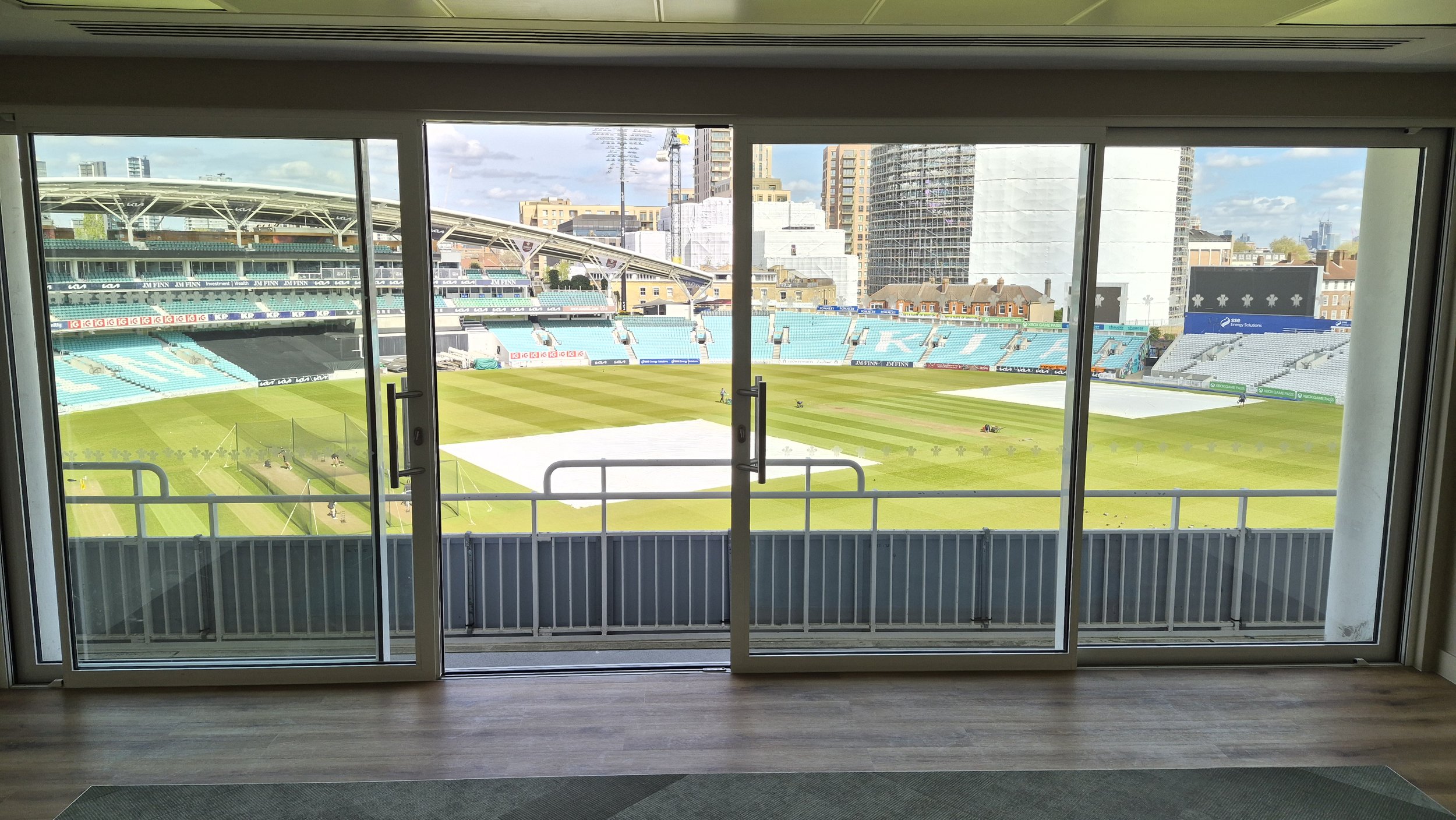 Indoor view overlooking a cricket stadium through glass doors, with a well-manicured field and seating sections, city buildings, and partly cloudy sky in the background.