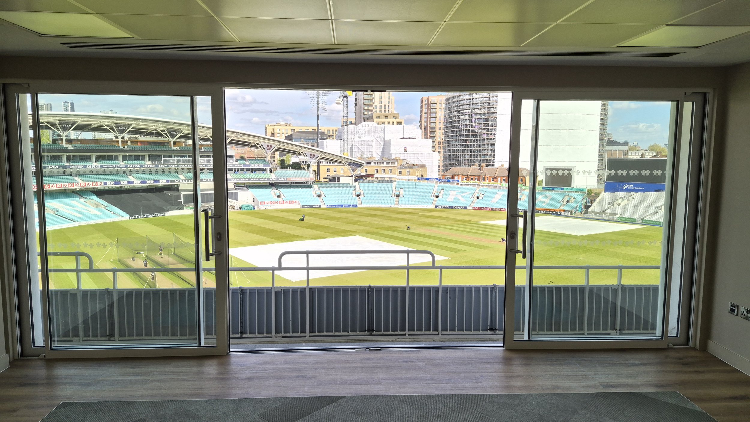 View of a cricket stadium from a room with glass sliding doors, showing green field, stands, and surrounding buildings in the background.