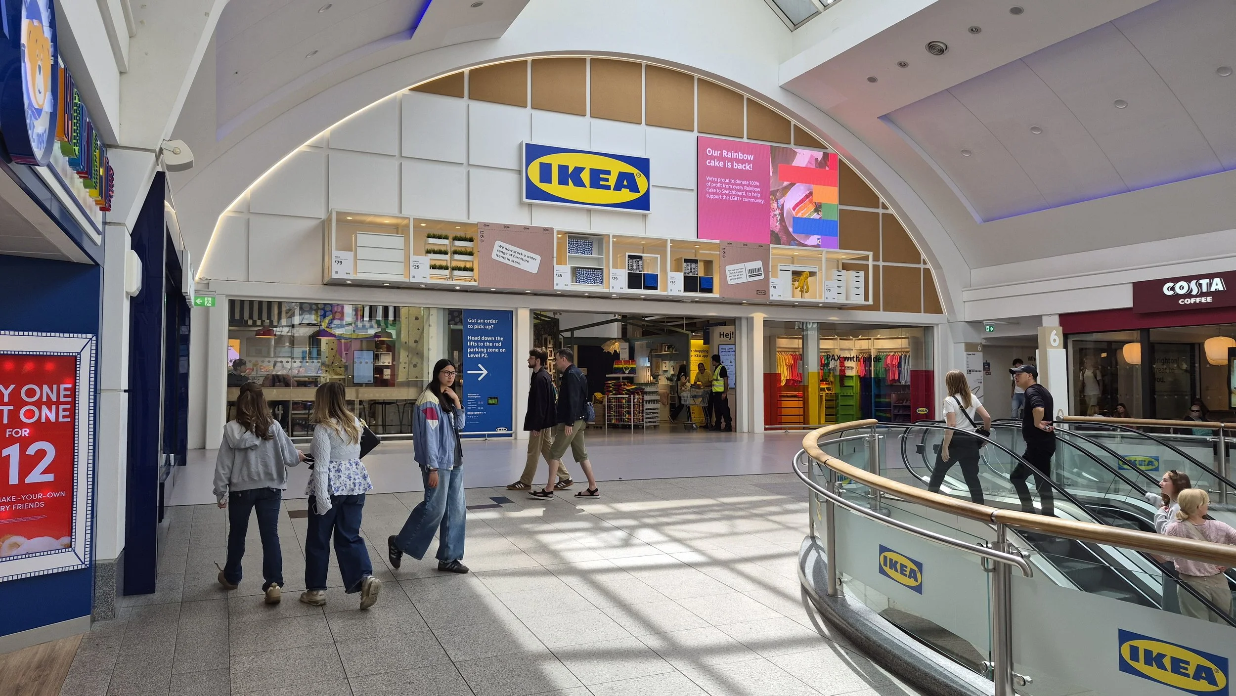 Inside a shopping mall with an IKEA store, digital screens, and people walking around, some using escalators.