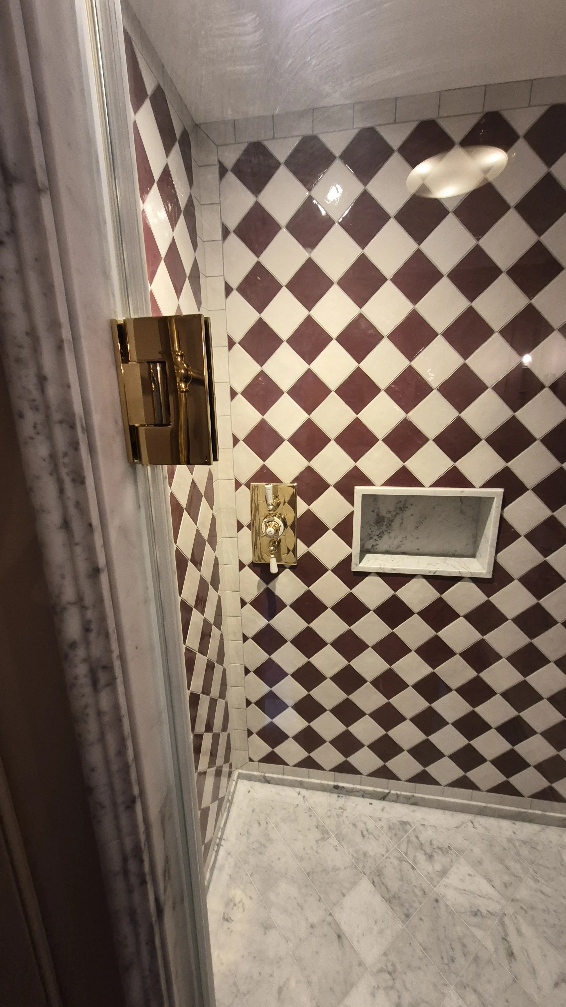 A bathroom shower area with a wall decorated in a red and white diamond pattern. There is a built-in marble shelf in the wall and a marble threshold at the entrance. The shower has a shiny brass faucet and fixtures.