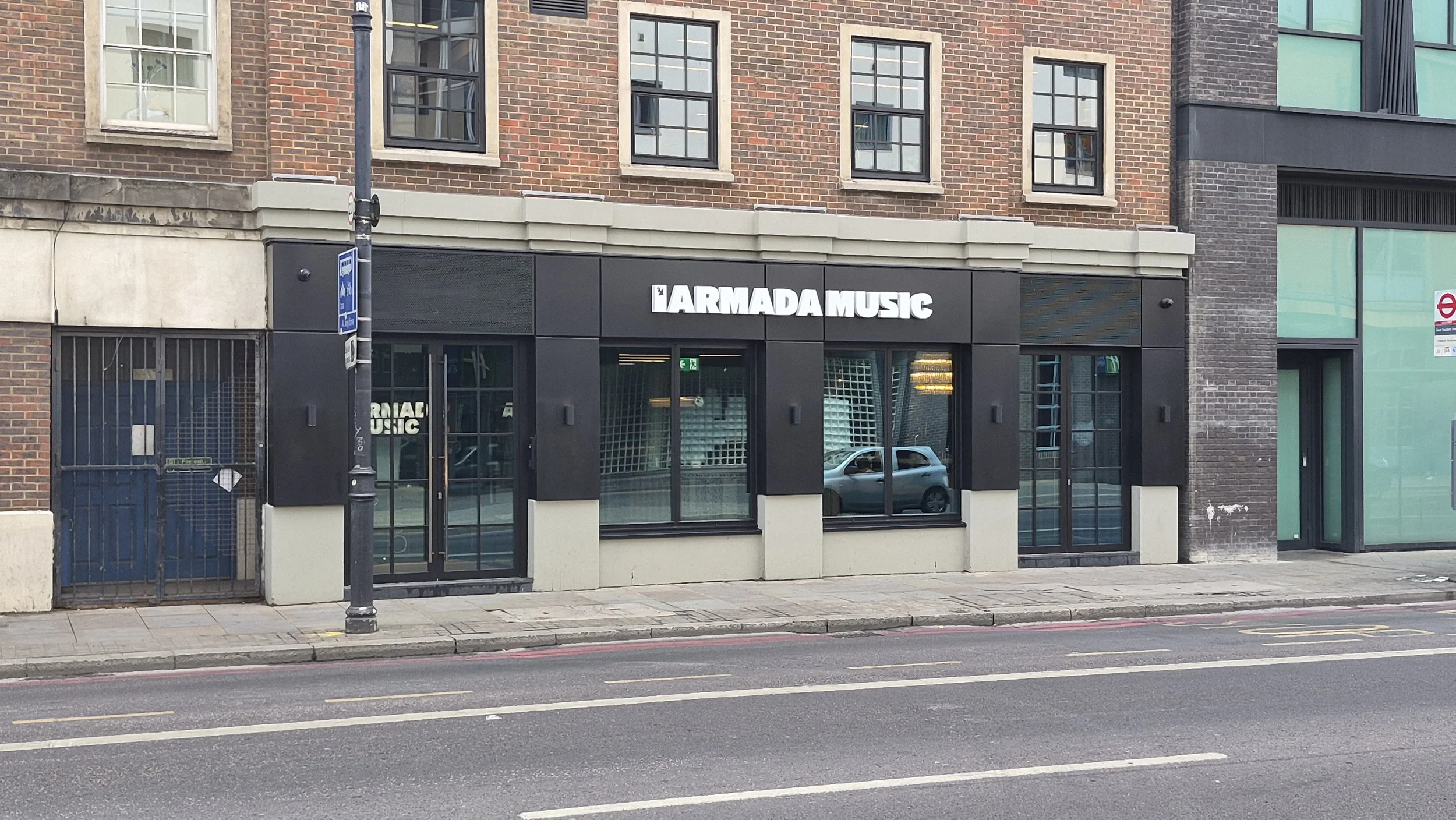 Storefront for Armada Music with large sign, glass windows, brick building, parked silver car visible inside through the windows, on a city street with a lamppost and bus stop sign.
