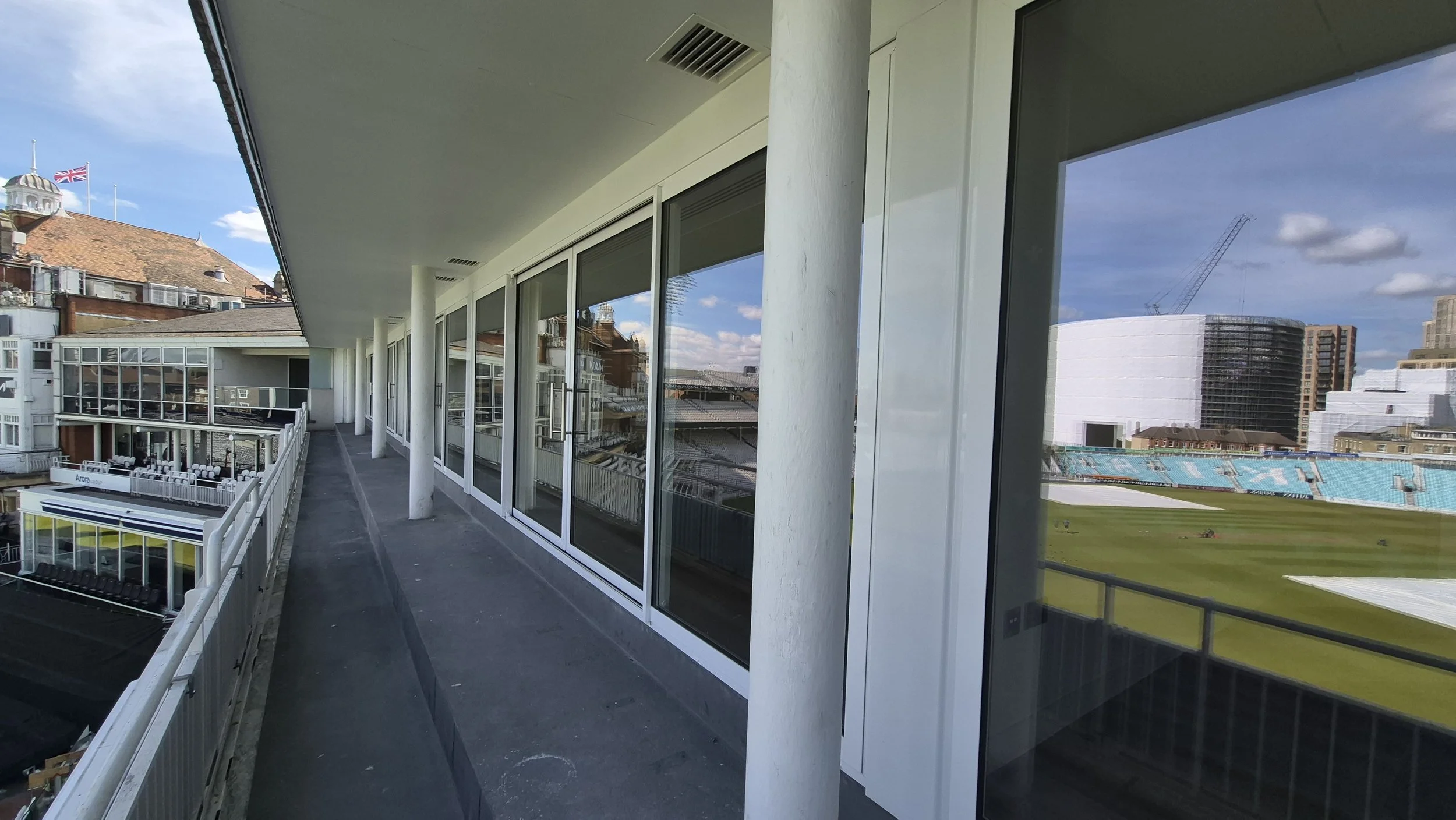 View from a balcony of a stadium, showing glass windows, white columns, and a cricket field with seating and structures in the background.