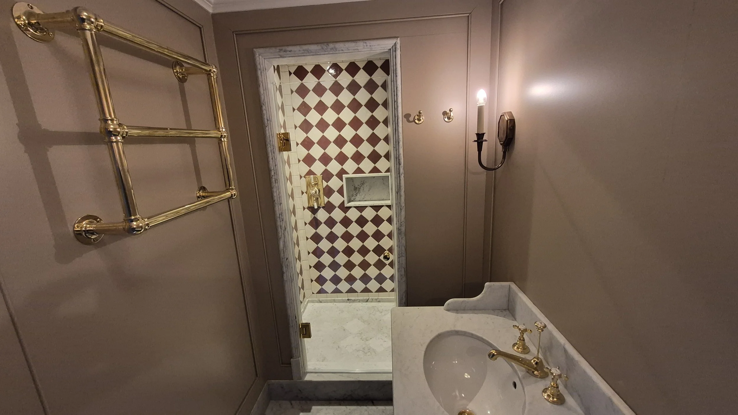 A small, elegant bathroom with dark walls, a marble sink with gold fixtures, a mirror, wall-mounted candle sconces, and a door with a gold handle leading to a room with checkered floor tiles.