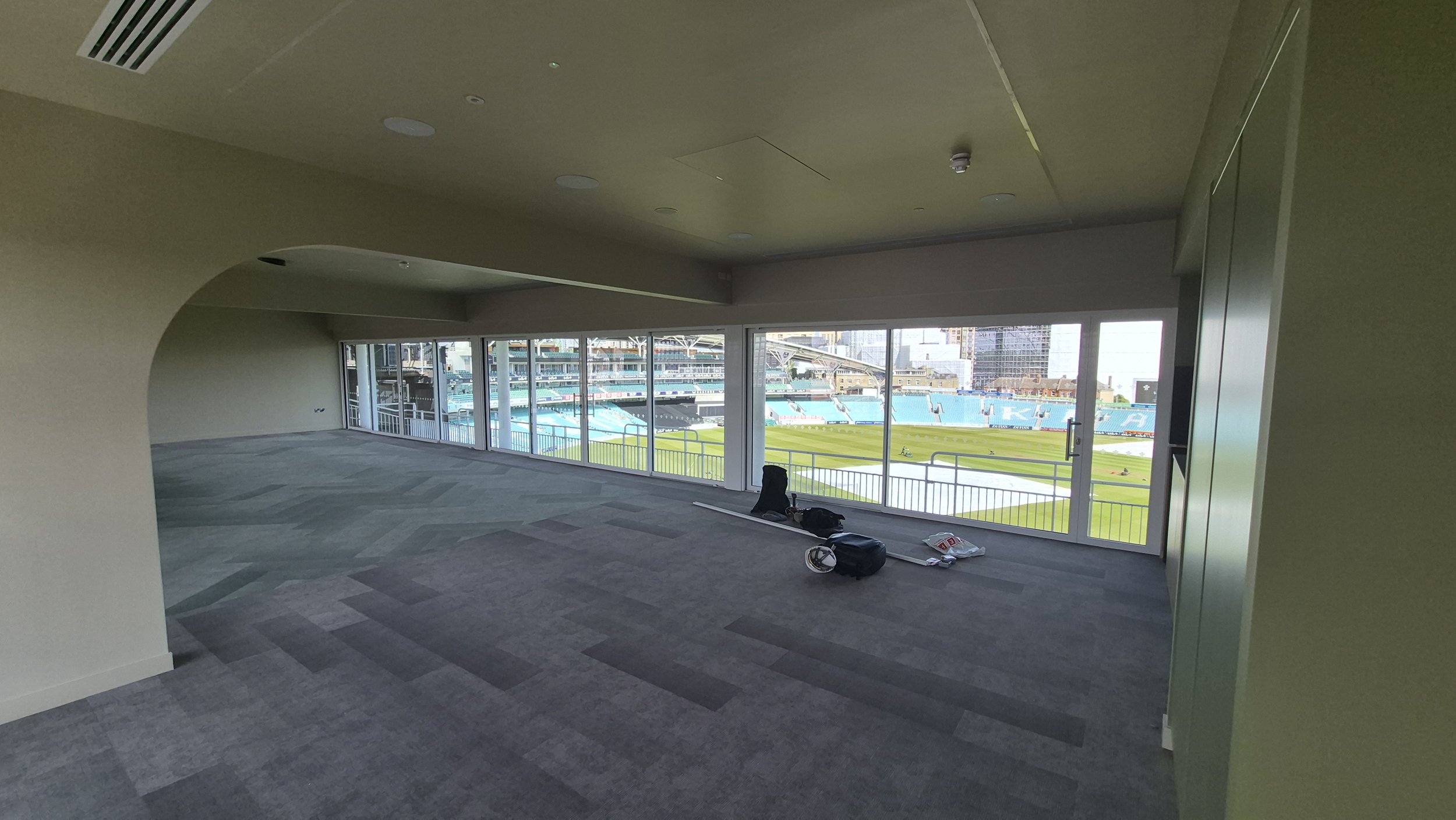 An empty indoor room with large windows overlooking a sports stadium with a grass field and seating. Items, such as bags and a helmet, are on the floor in the room.