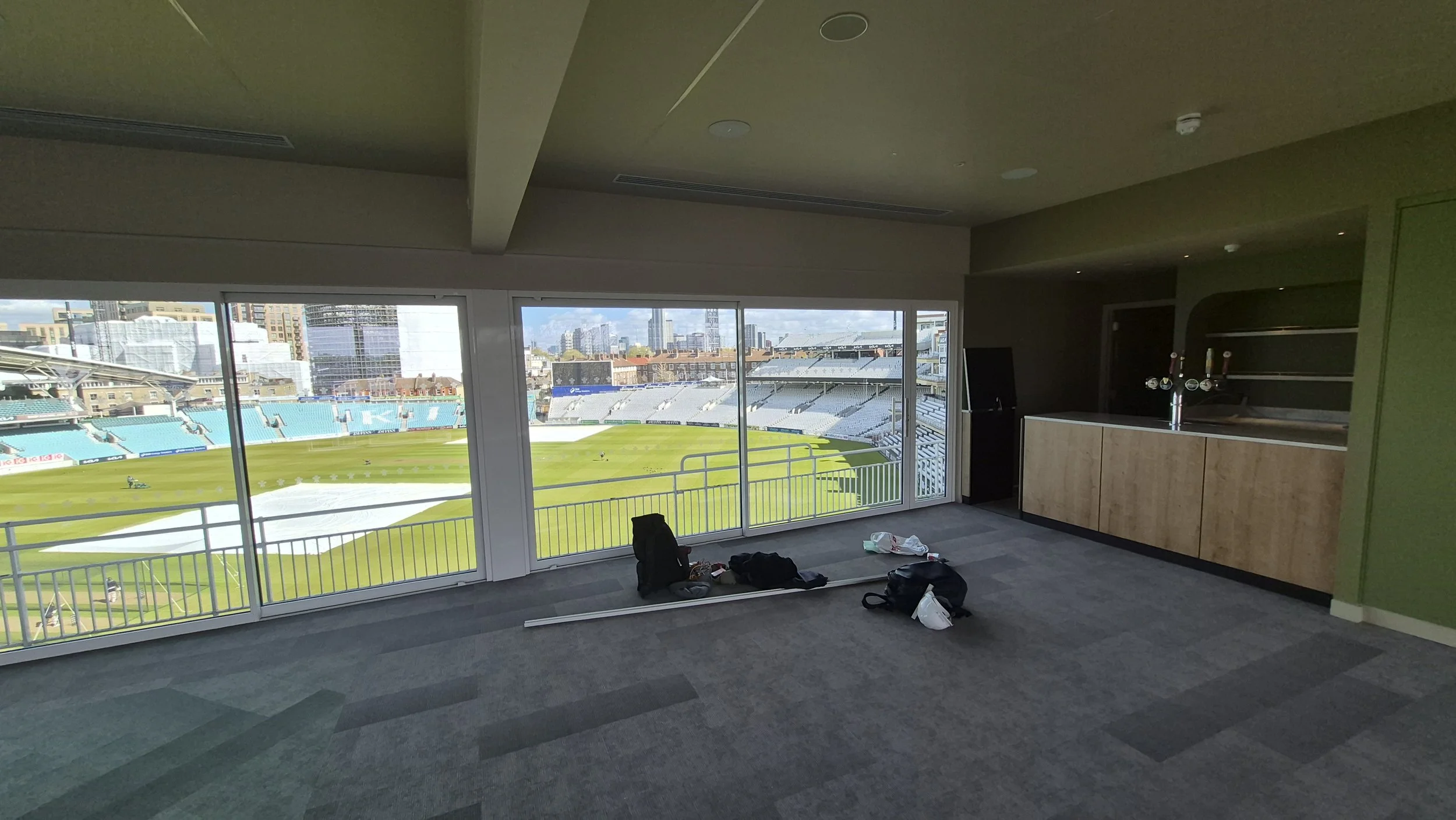 Interior of a room with large windows overlooking a cricket stadium, with objects and bags on the floor.