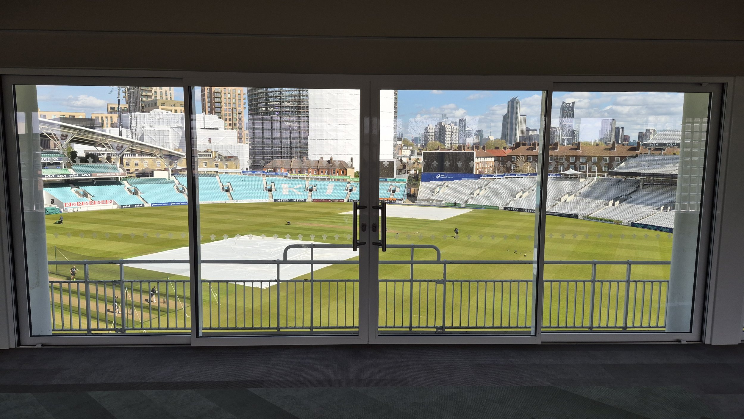 View of a cricket stadium through glass doors, with the field, seating area, and city skyline in the background.