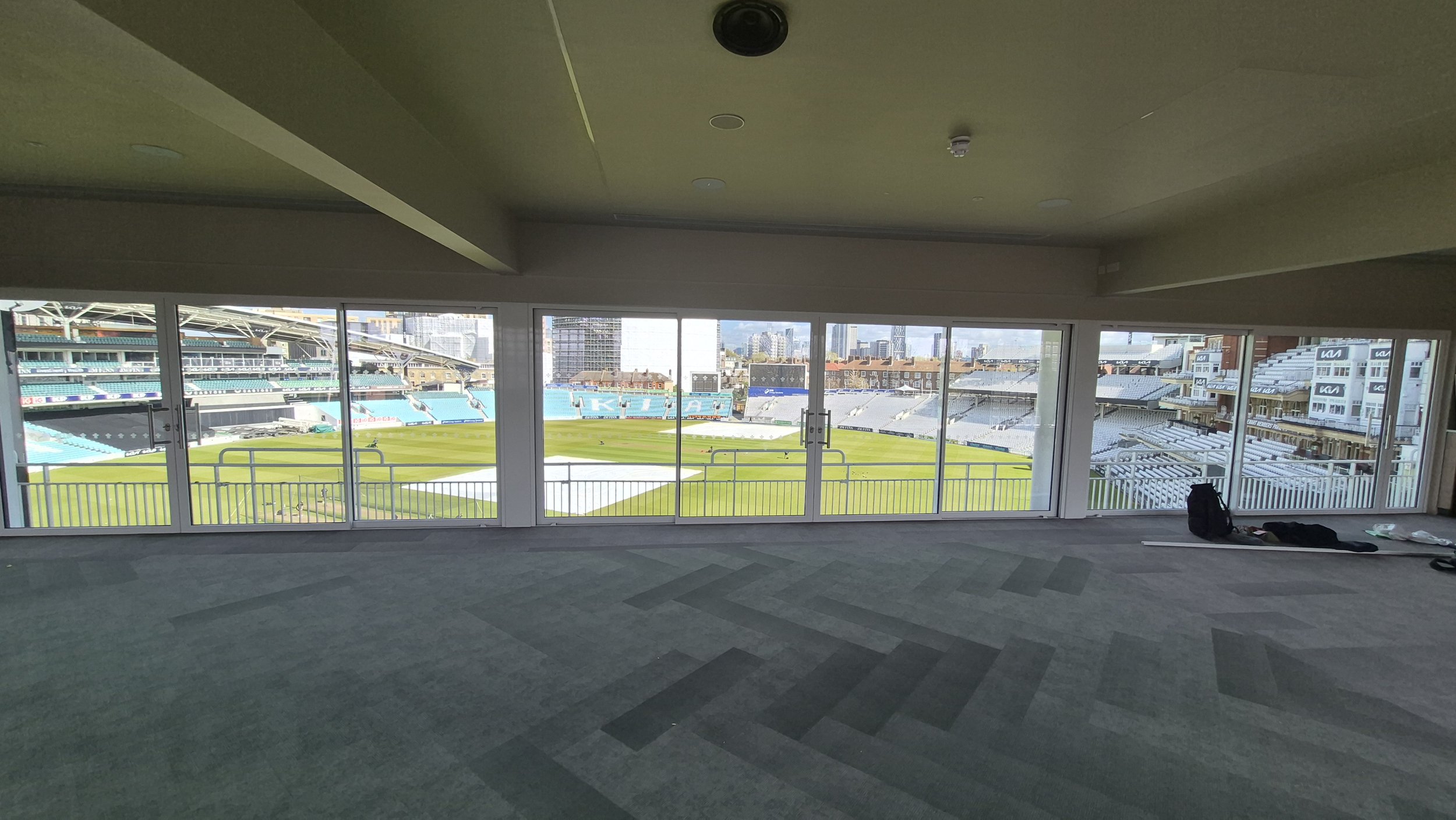 Interior view of a room with large glass doors overlooking a cricket stadium with a field, seating areas, and cityscape in the background.