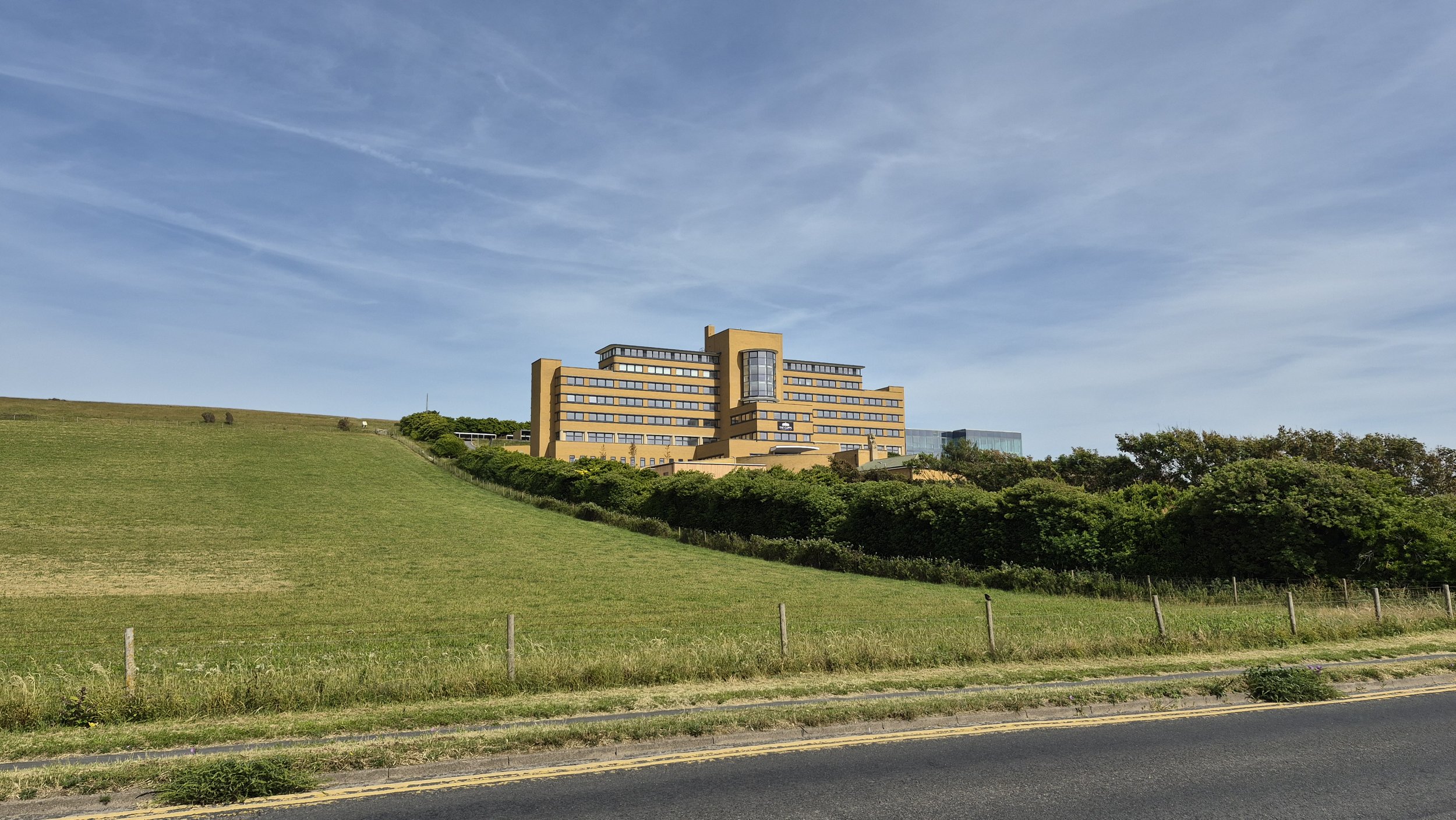 A large yellow-brick building on a hill surrounded by green trees and grass, with a road in the foreground and a blue sky above.