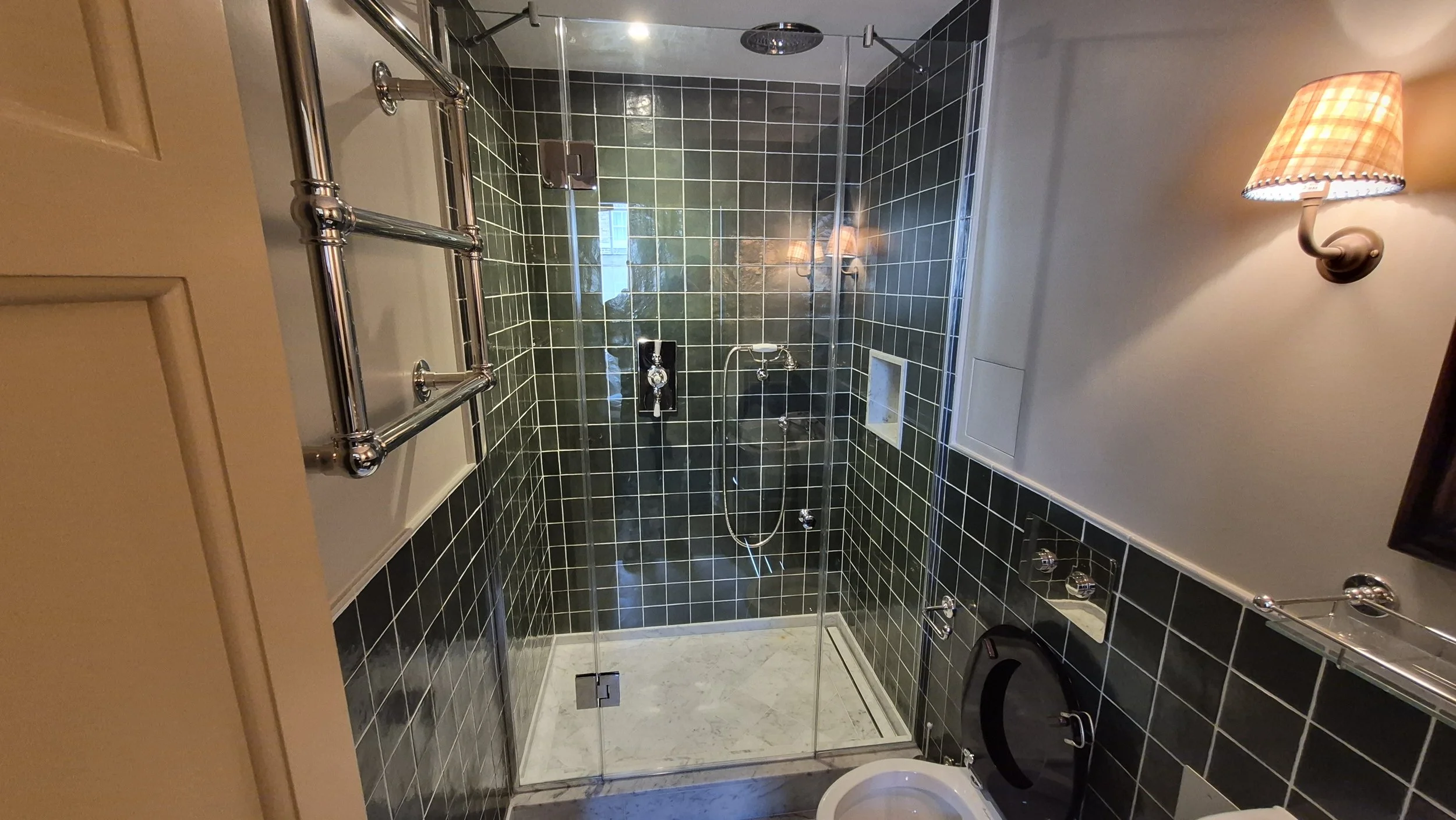 A small tiled bathroom with a glass shower enclosure, a wall-mounted lamp with a beige lampshade, and a toilet. The shower has black tiles on the walls and a small niche for toiletries.