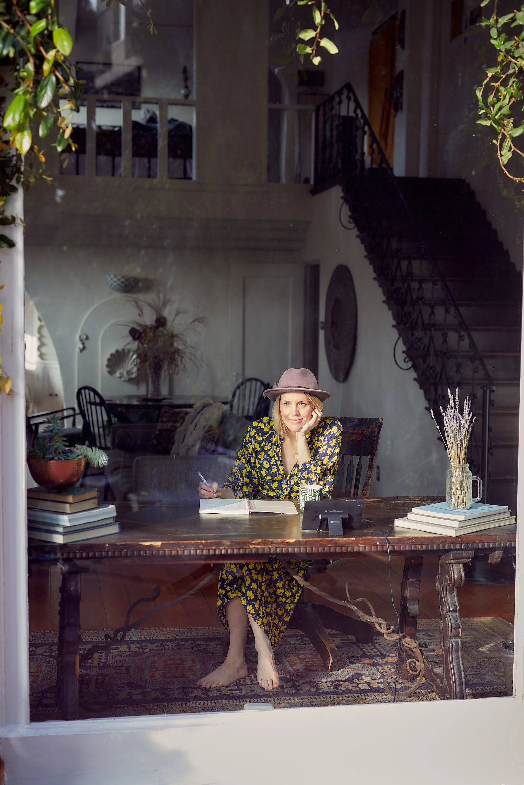 Woman sitting at a wooden desk with a notebook, pen, and mug, seen through a glass window. The woman is wearing a patterned dress and a hat, resting her head on her hand. The room behind her has a staircase, books, and decorative plants.