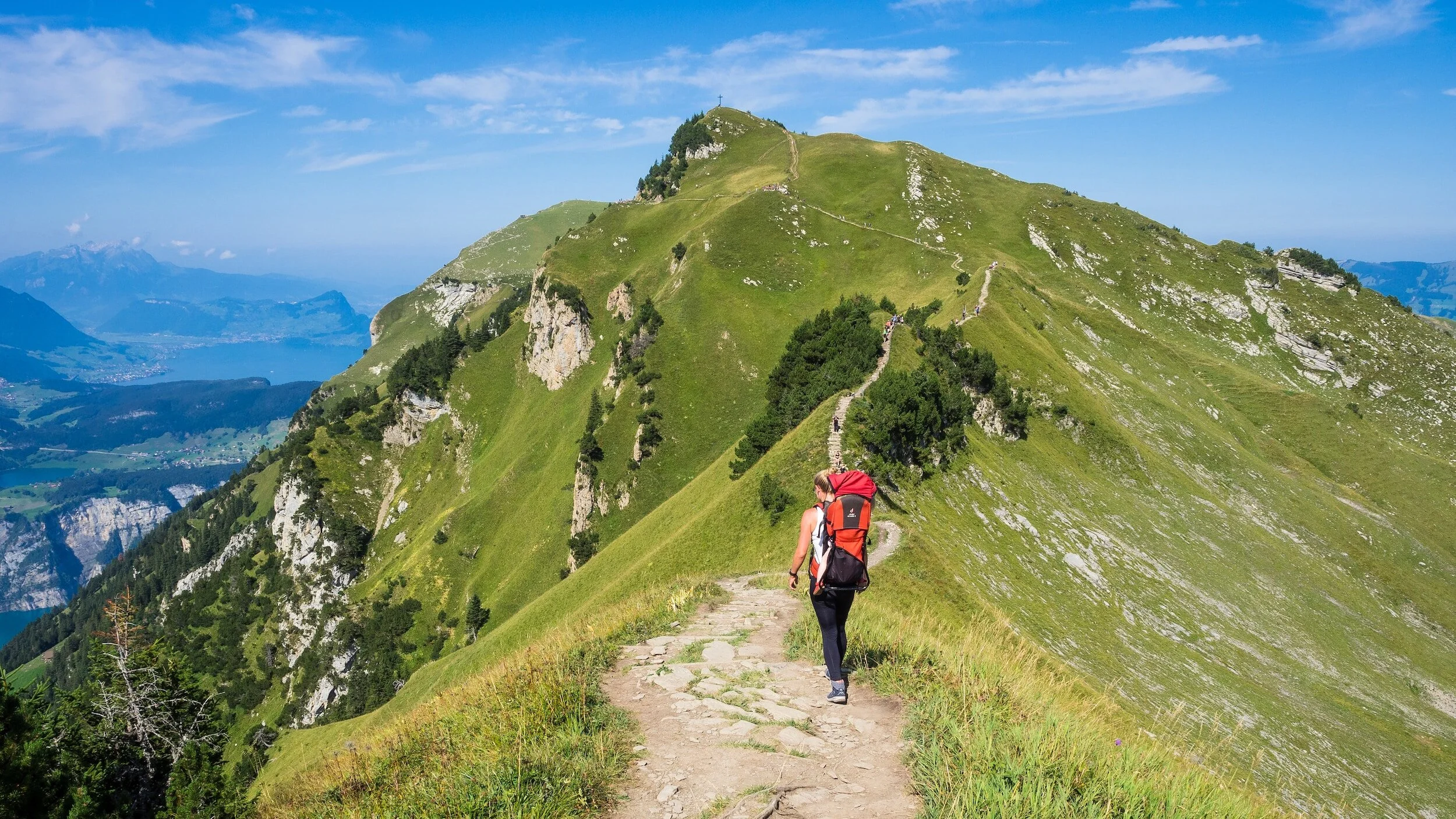 Hiker walking on a mountain trail with green grass and shrubs, overlooking distant lakes and mountains under blue sky