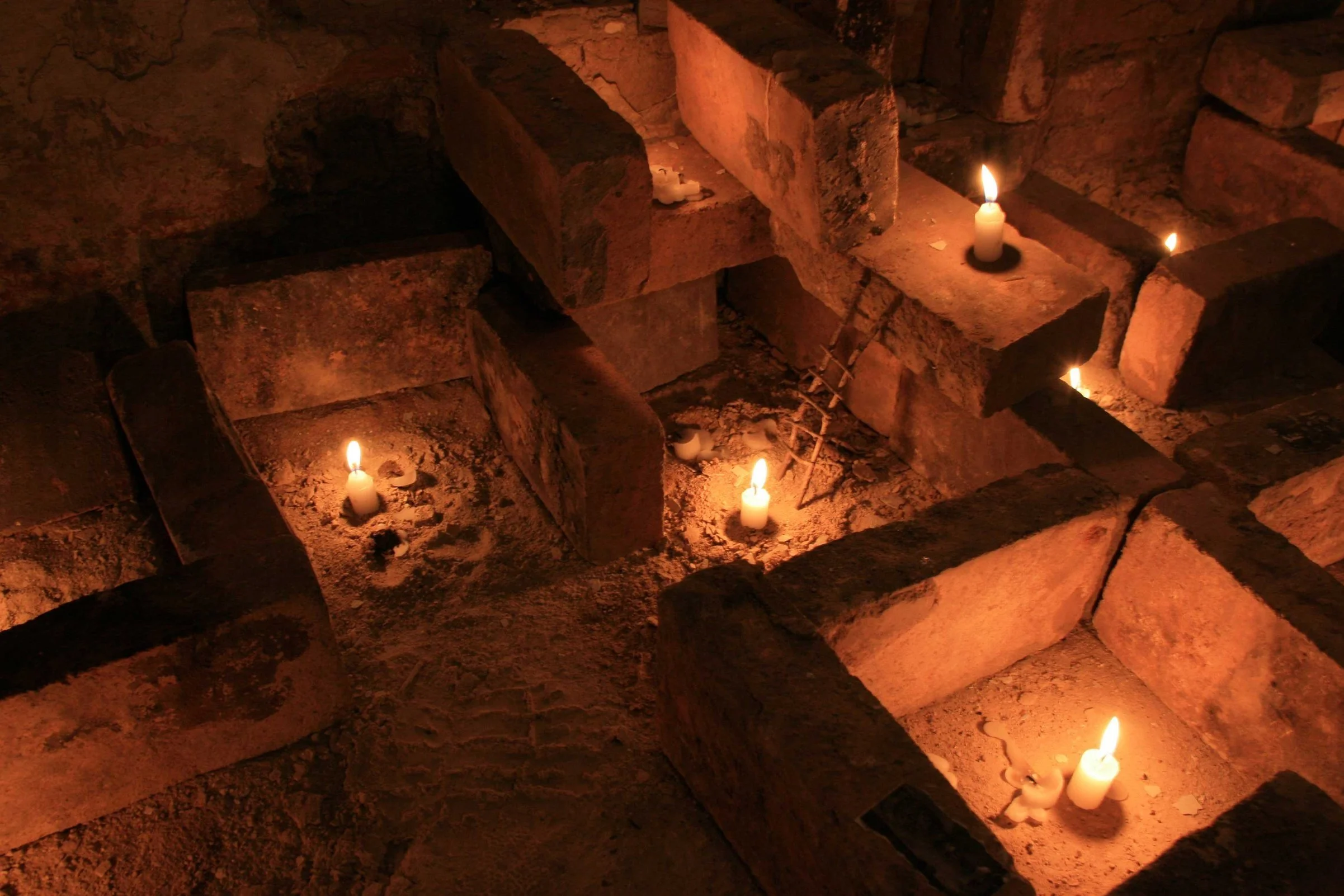 Velas encendidas en un lugar con paredes de ladrillos y una tumba o sepulcro, dando un ambiente sombrío y solemne.
