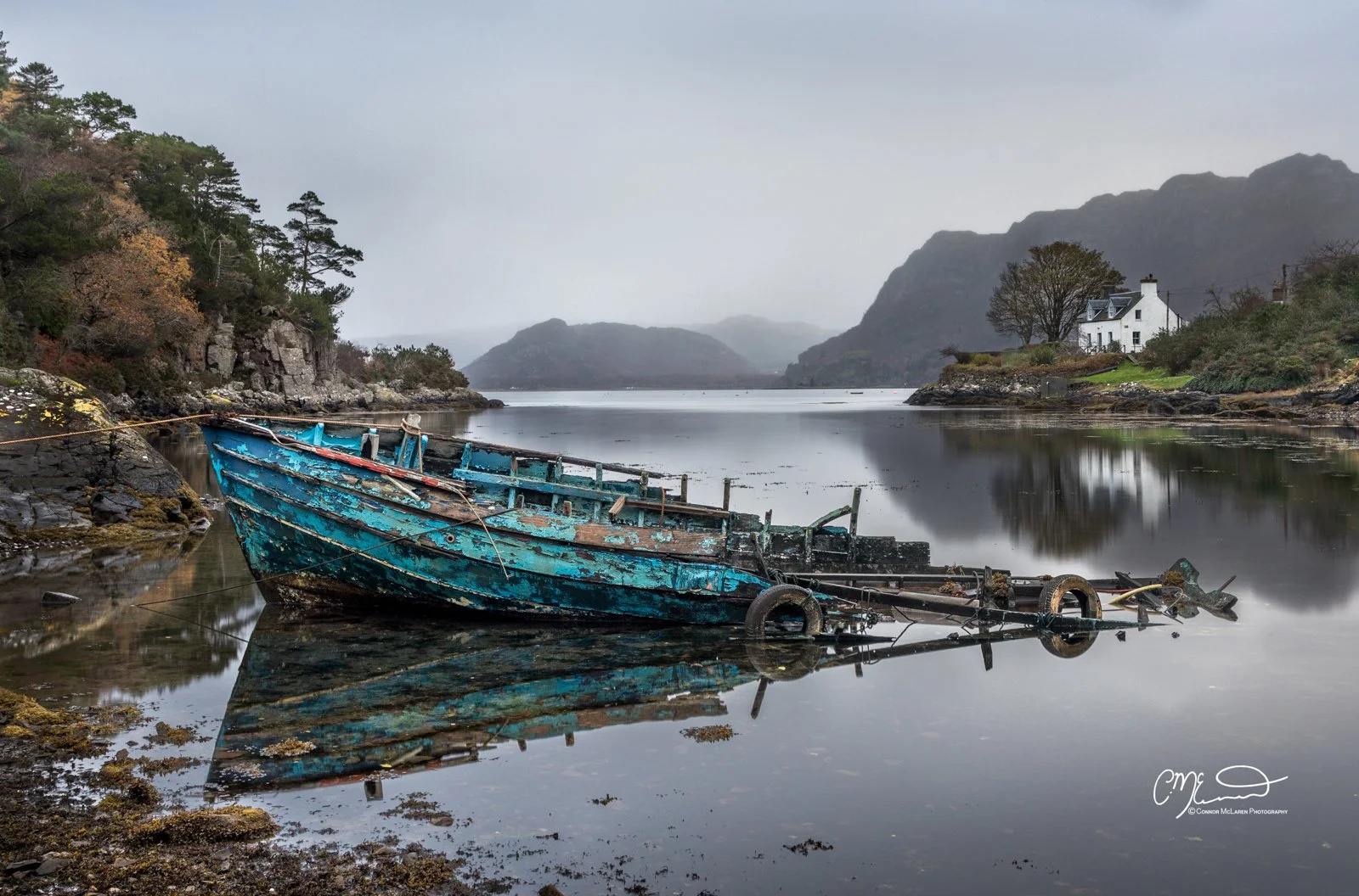 Plockton, western highlands scotland. A weathered, blue, abandoned boat partially submerged and tilted at an angle in calm water near the shore of a serene lake or river, with a foggy, mountainous landscape and a white house in the background.