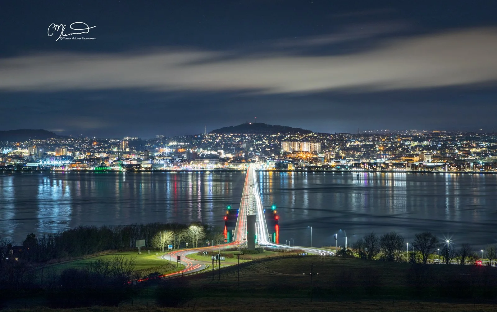Dundee, North East Scotland. Nighttime cityscape with a long illuminated bridge over a body of water, reflecting city lights, with a hill in the background and a partly cloudy sky.
