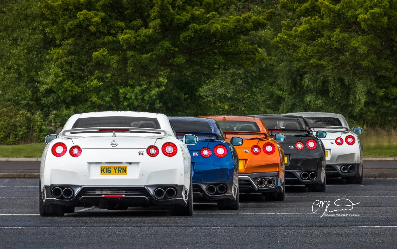 Four Nissan GT-R cars in white, blue, orange, and black, parked in a line on an empty parking lot with green trees in the background.