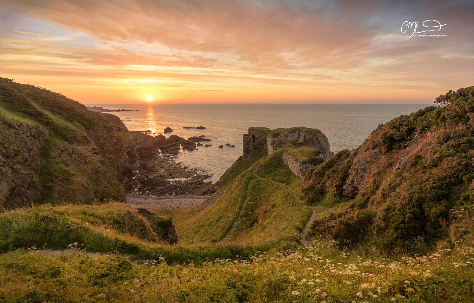 Findlater Castle, North East Scotland. Sunset over a coastal landscape with grassy hills, rocky cliffs, and an old stone ruins by the sea.