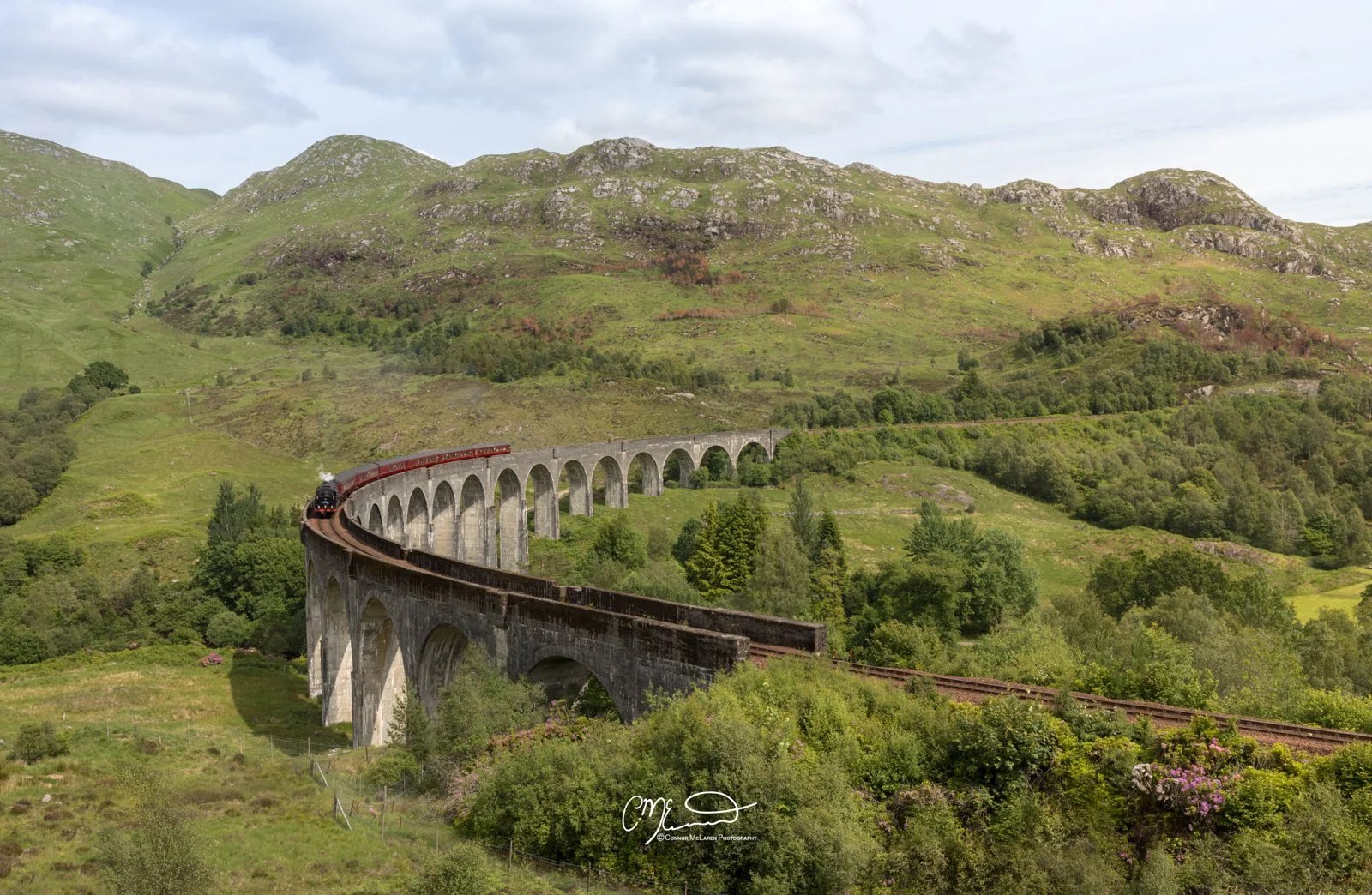 Bucket list locations, Scotland, Glenfinnan viaduct, jacobite steam train, harry potter train. A vintage steam train traveling on a curved stone viaduct bridge through a lush green valley with rolling hills and trees.